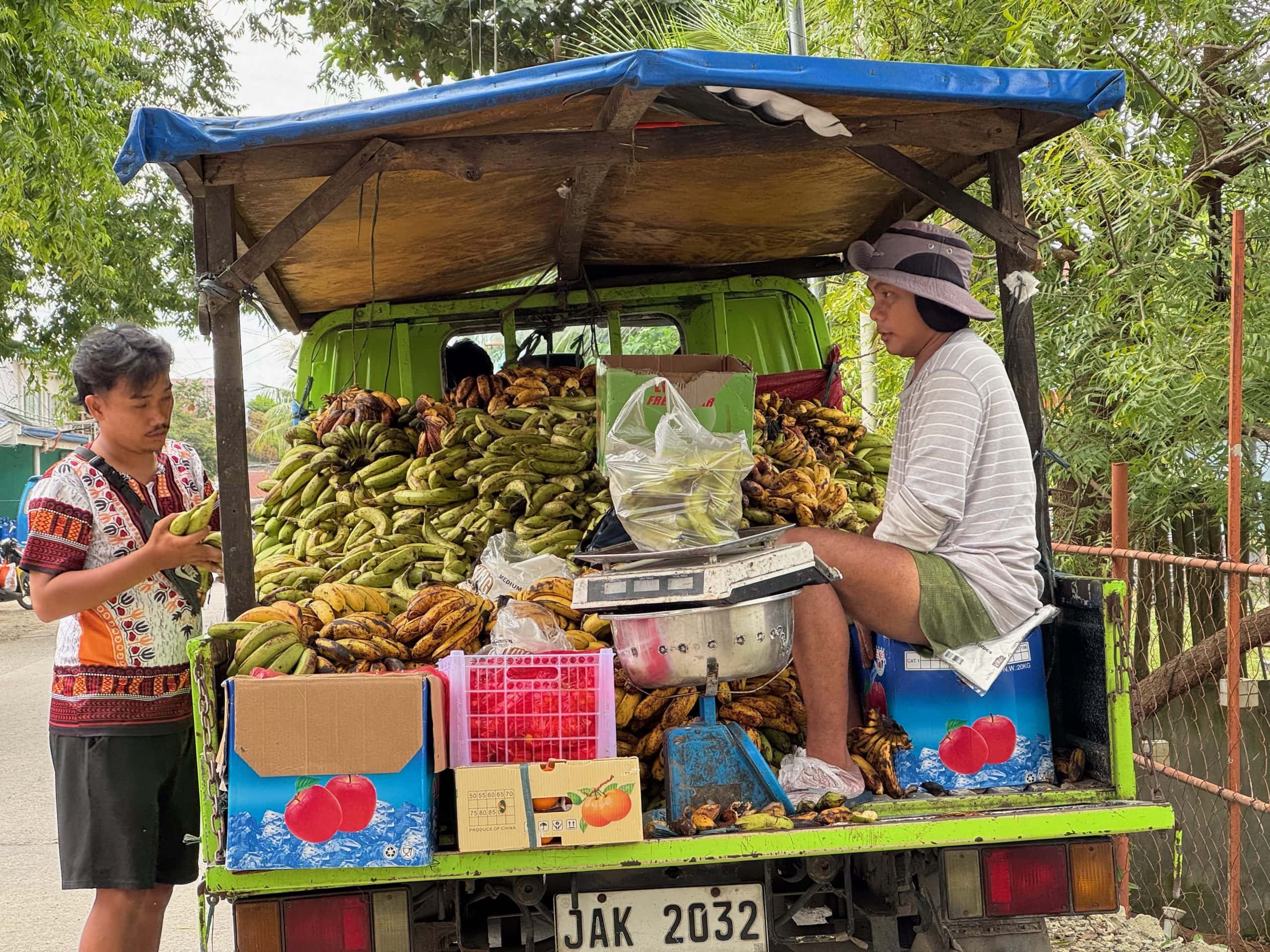 An image of a truck filled with bananas and a salesman selling bananas to the public.