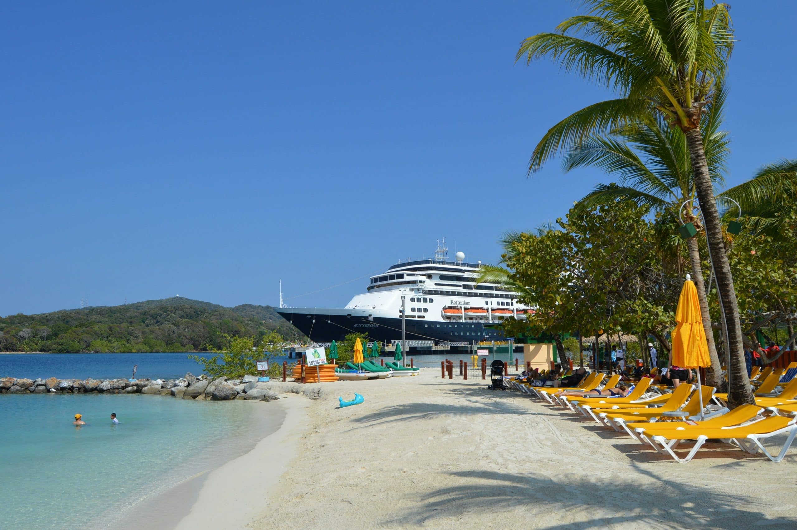 Palm-lined beach and clear ocean water in Roatan, Honduras under bright blue skies with a Roatan cruise ship in the background.