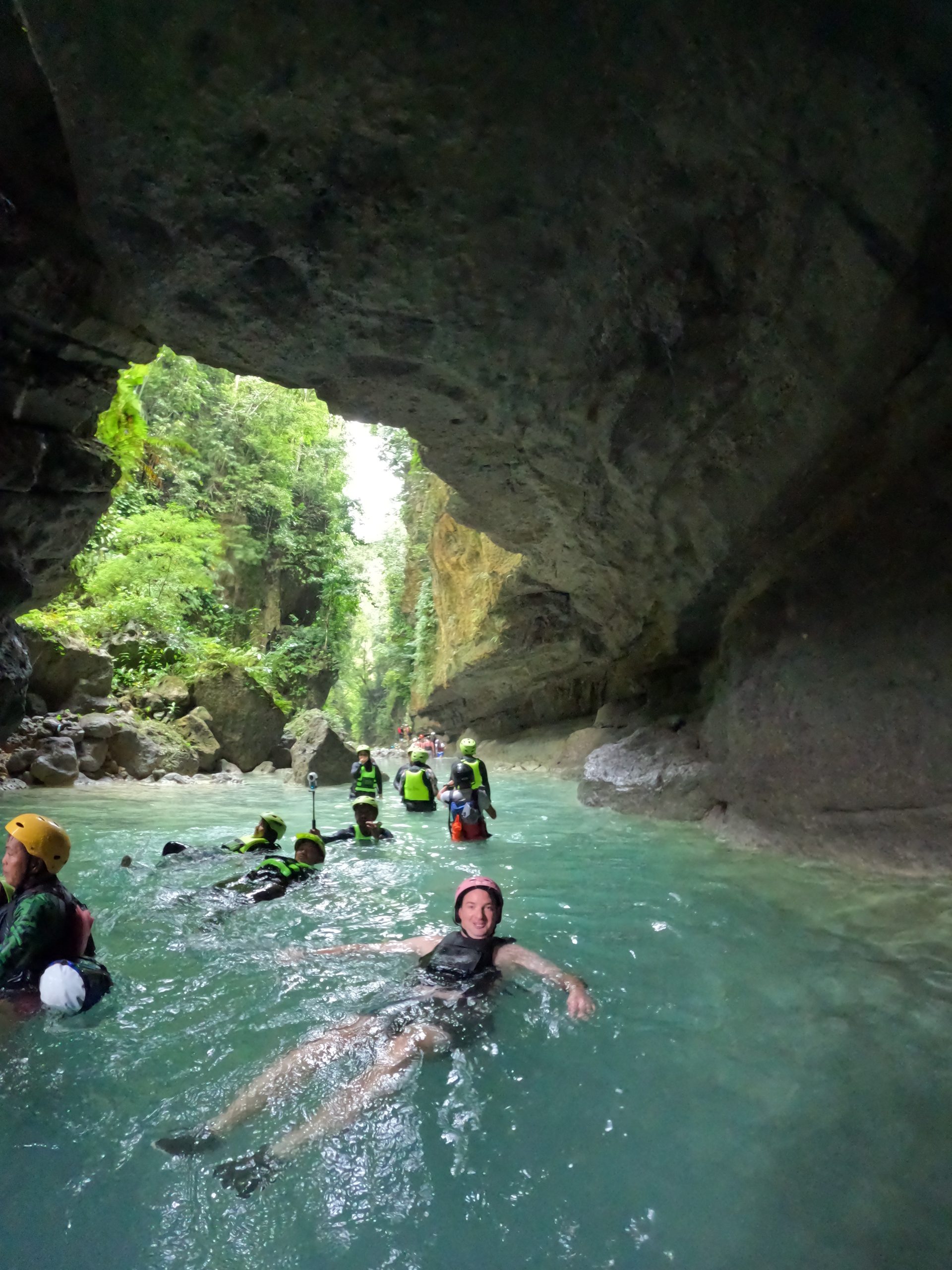 Tour group swimming through a cave opening at Kawasan Falls, surrounded by turquoise water and jungle cliffs — part of the Moalboal canyoneering experience.
