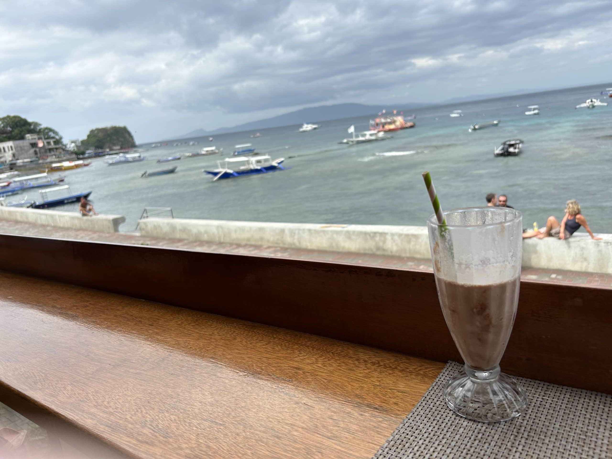 View from the restaurant at Cap’n Gregg’s Dive Resort in Sabang, Puerto Galera, showing a milkshake on the counter and boats anchored in the bay under cloudy skies.