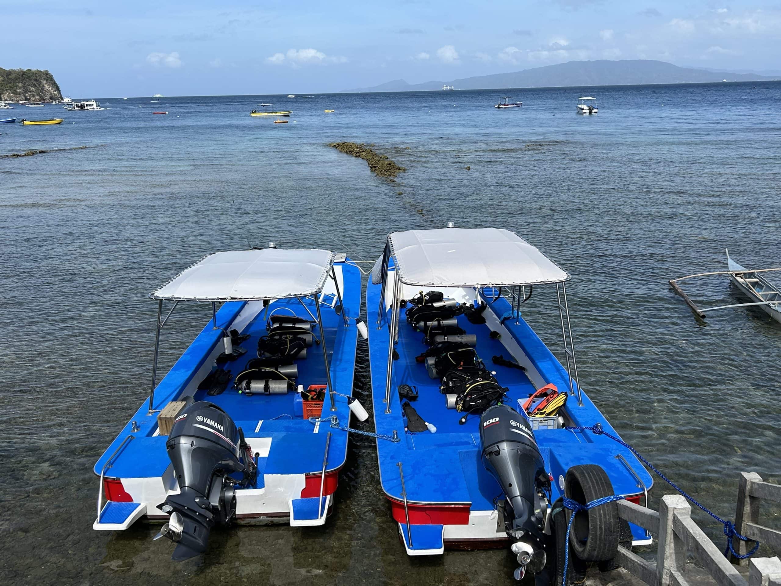 Twin blue-and-white dive boats docked in shallow water at Sabang Beach, ready for scuba diving trips in Puerto Galera.