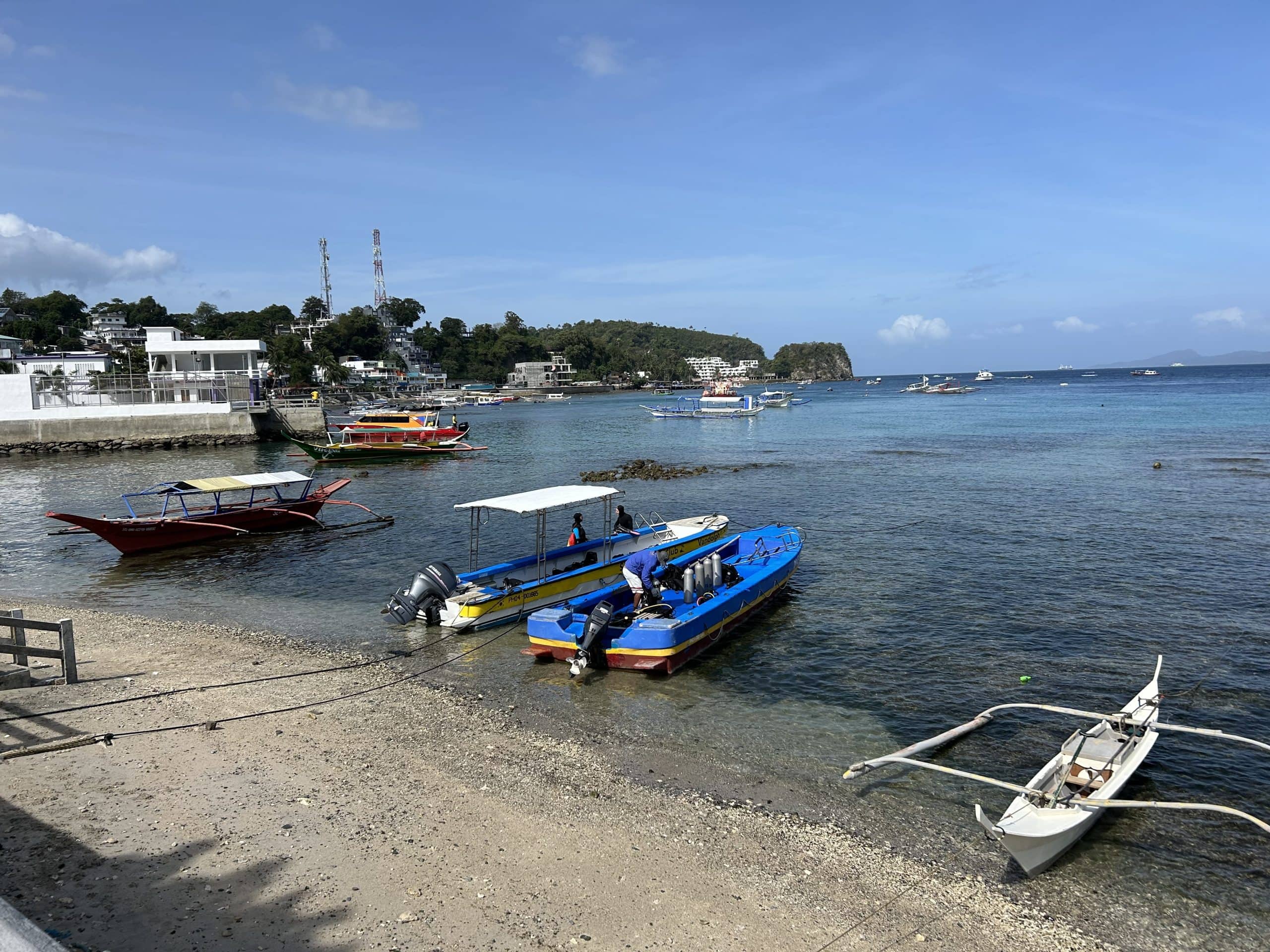 Boats anchored along the sandy shoreline of Sabang Beach, with resorts and hills visible in the background under a clear sky.
