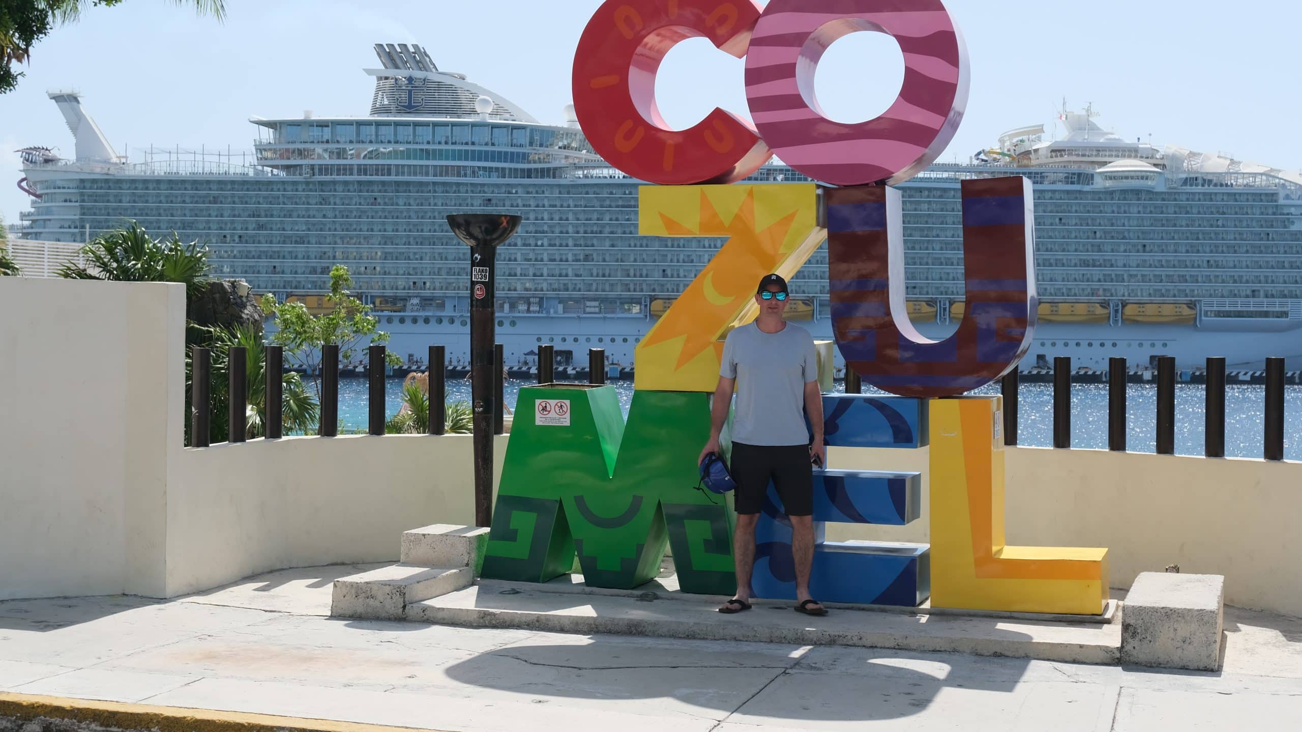 Tourist posing in front of the colourful Cozumel sign with a massive cruise ship docked in the background on a sunny day in Mexico.