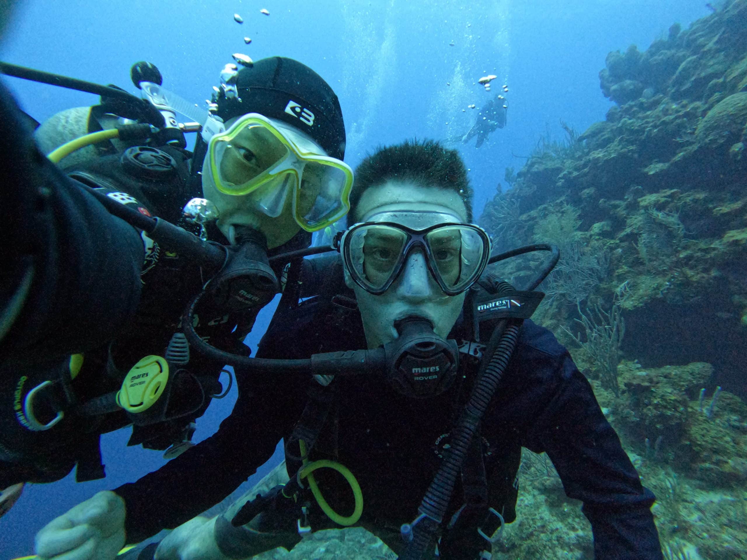 Two scuba divers pose underwater beside a vibrant coral wall in Cozumel, Mexico, capturing a selfie during a drift dive in clear blue Caribbean waters.