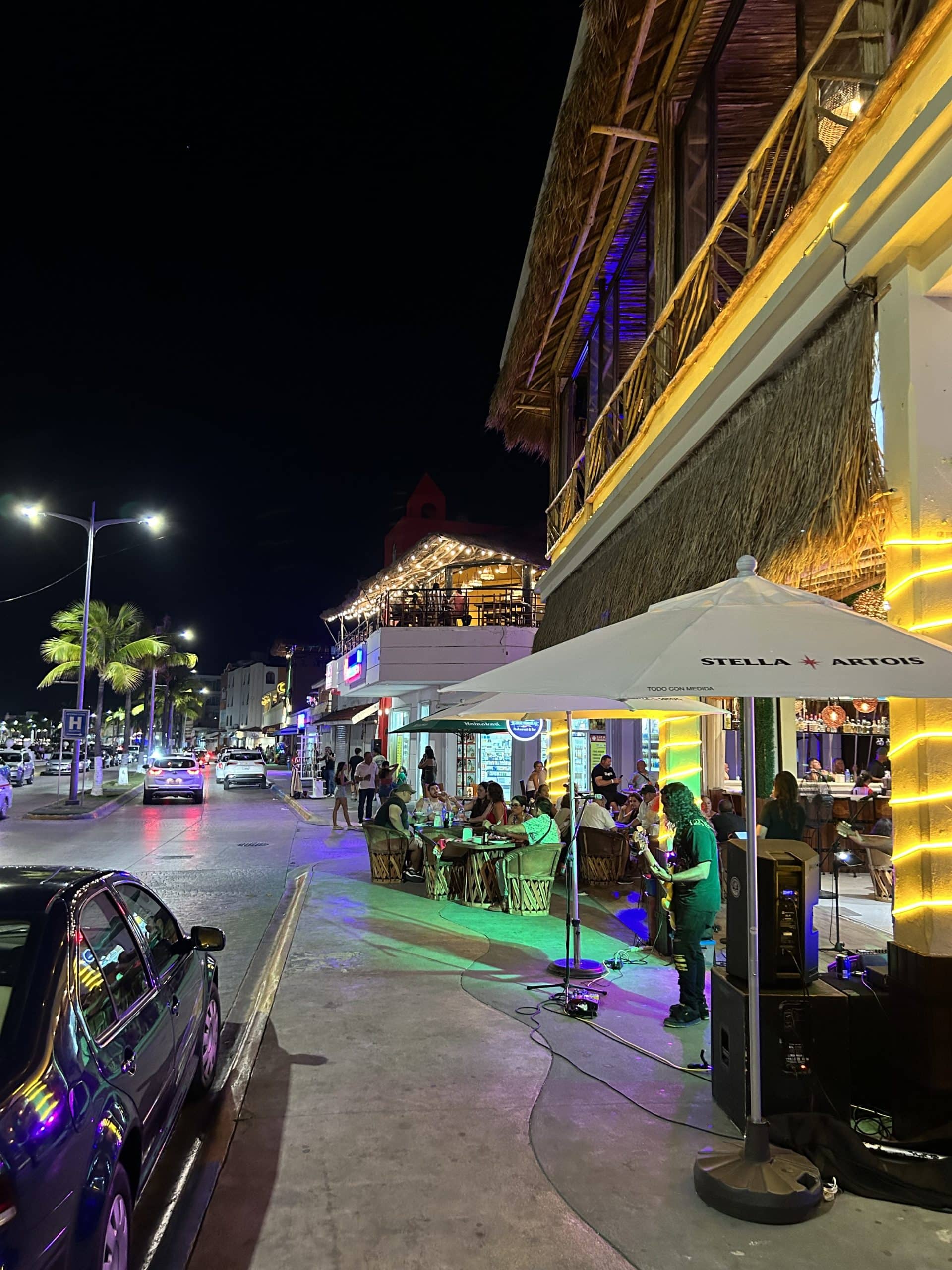 Vibrant Cozumel nightlife scene along the waterfront with live music, outdoor dining, and palm-lined streets lit up at night.