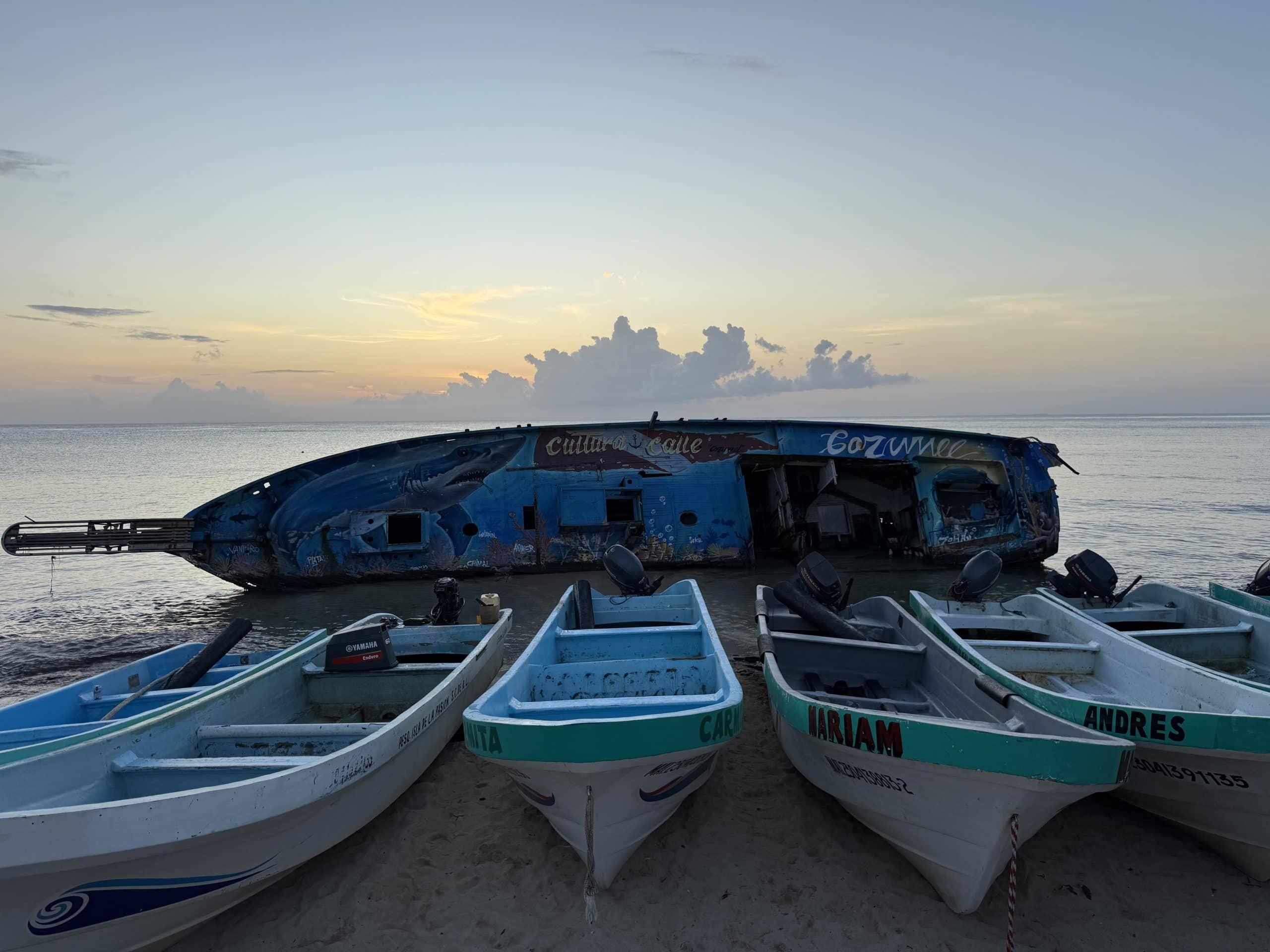 Colourful fishing boats rest on the beach at sunrise in Cozumel, with a painted shipwreck in the background and calm ocean waters under a glowing sky.