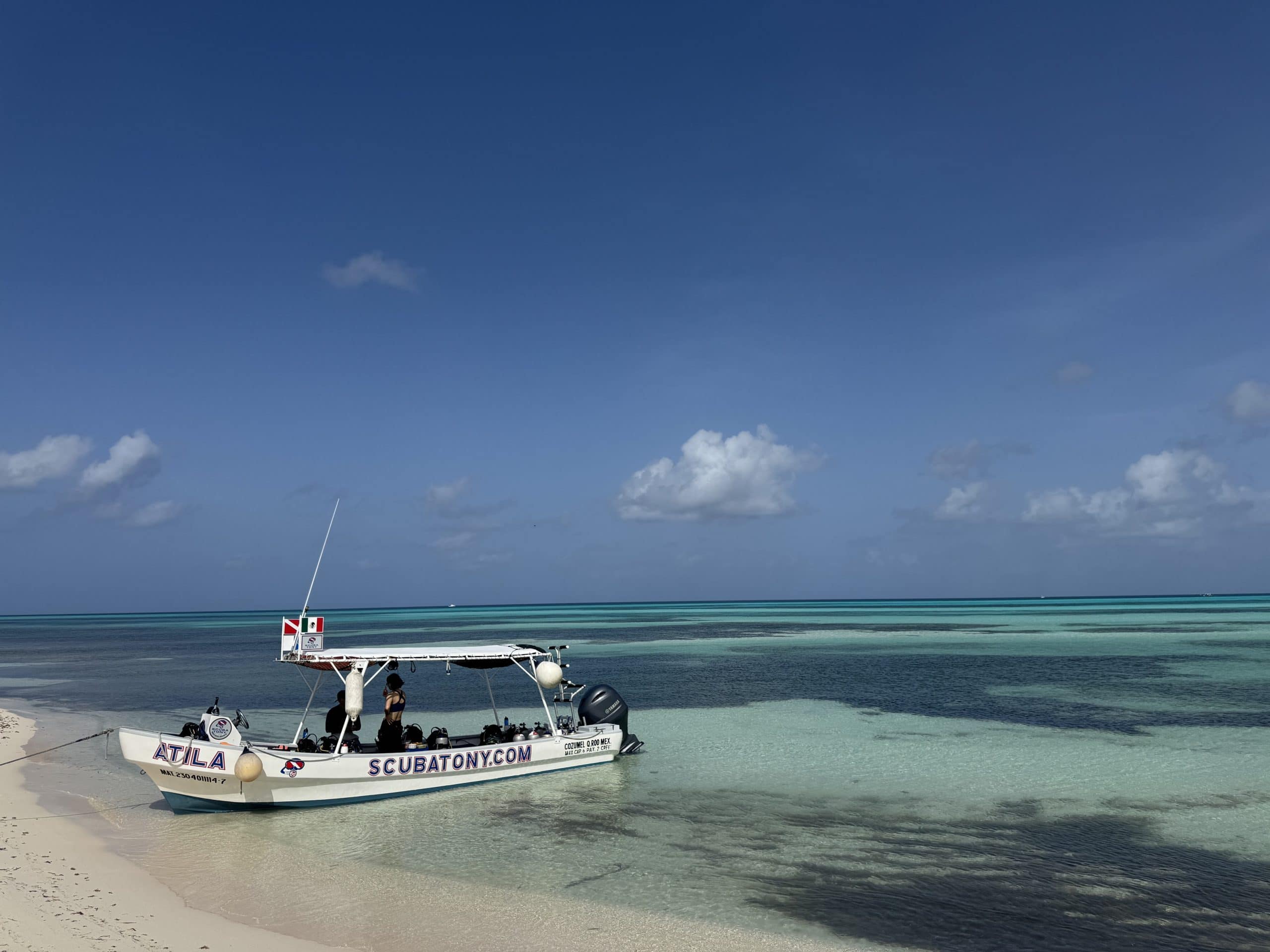 ScubaTony dive boat anchored on a shallow white sand beach in Cozumel, Mexico, with turquoise Caribbean waters and a clear blue sky in the background.