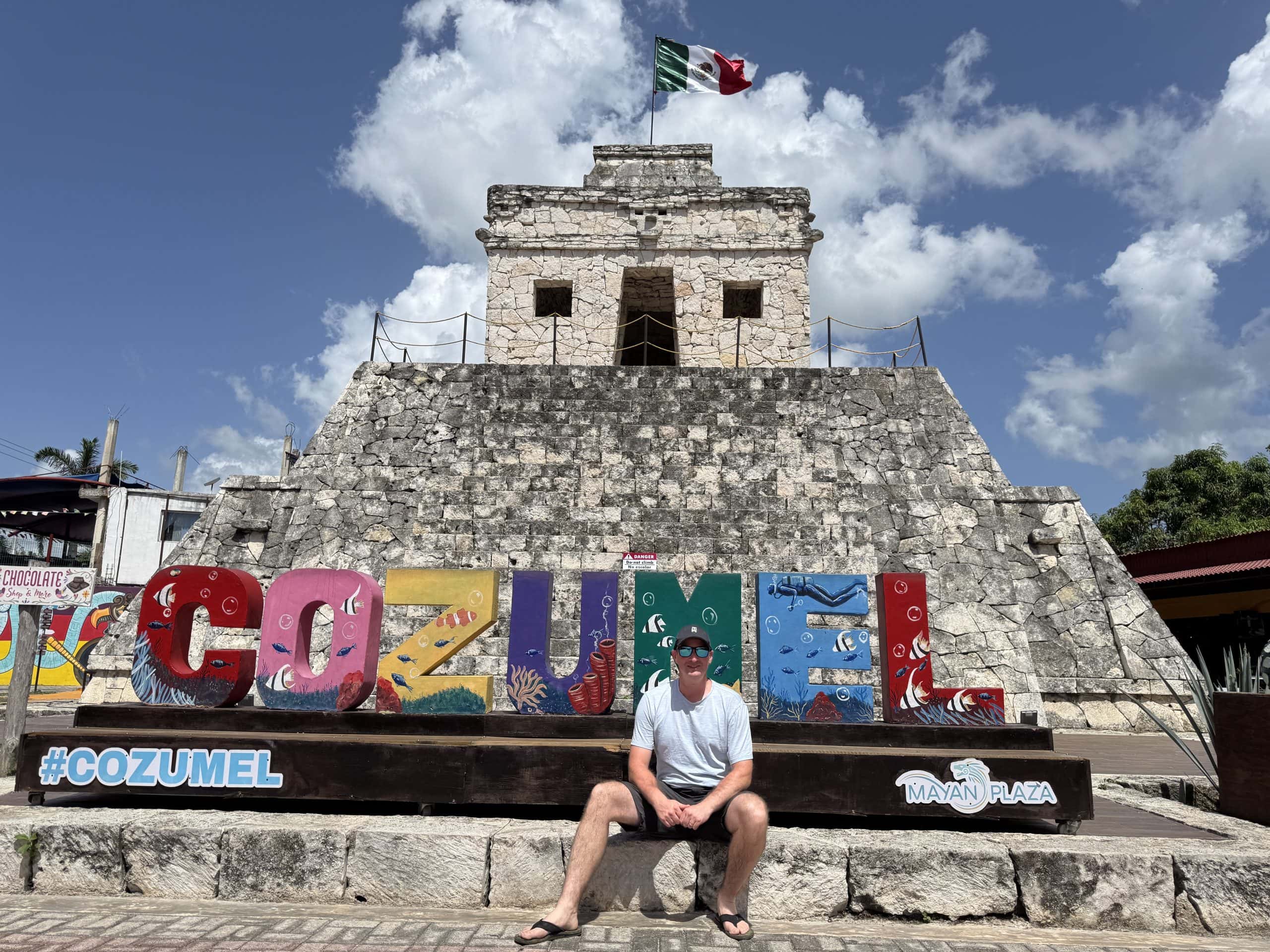 A traveller sits in front of the colourful Cozumel sign at Mayan Plaza with a replica Mayan temple behind him and the Mexican flag flying above, under a bright blue sky.
