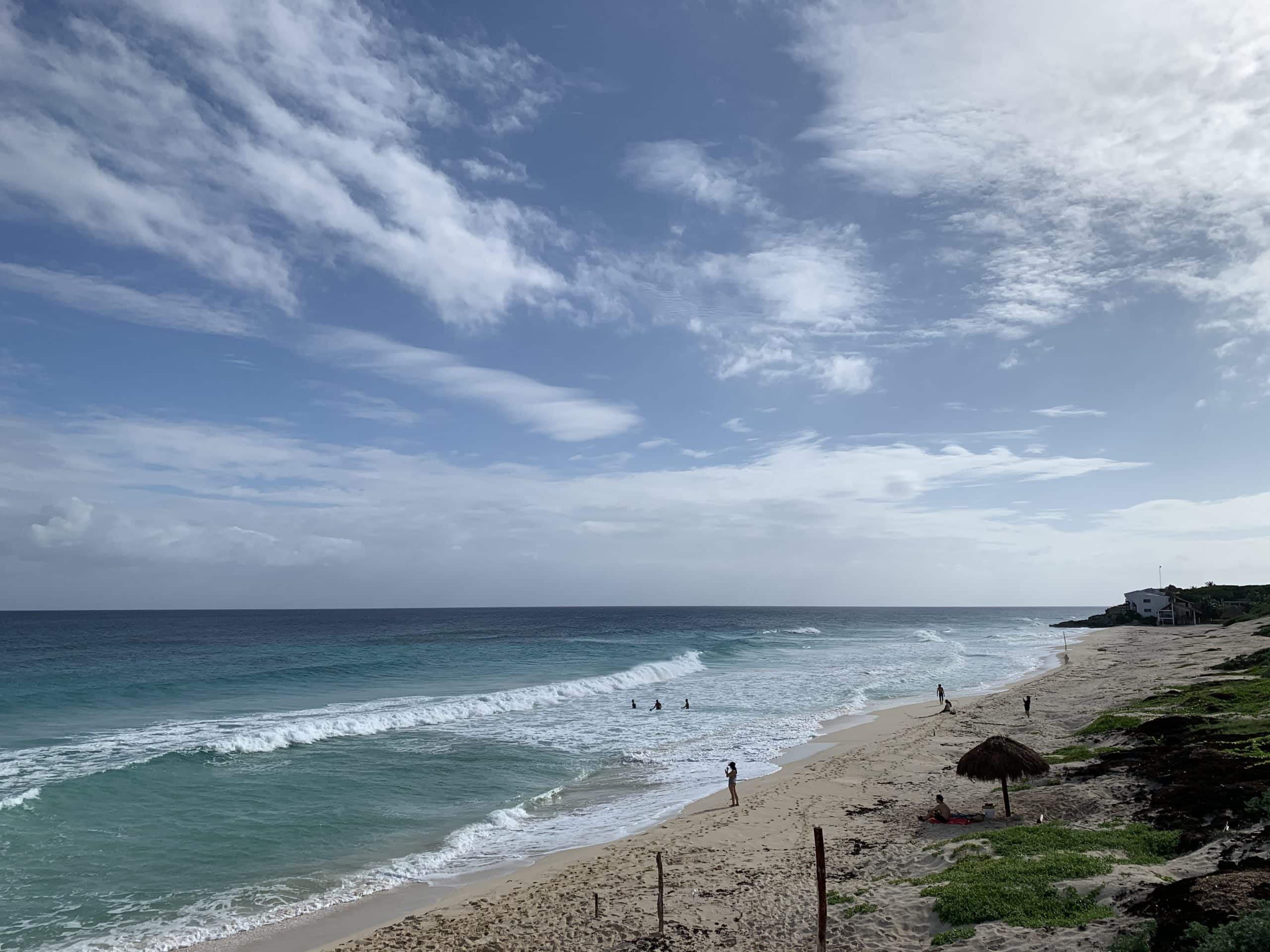 Peaceful beach on the eastern coast of Cozumel with turquoise waves, soft sand, and a few swimmers enjoying the surf under a bright blue sky.