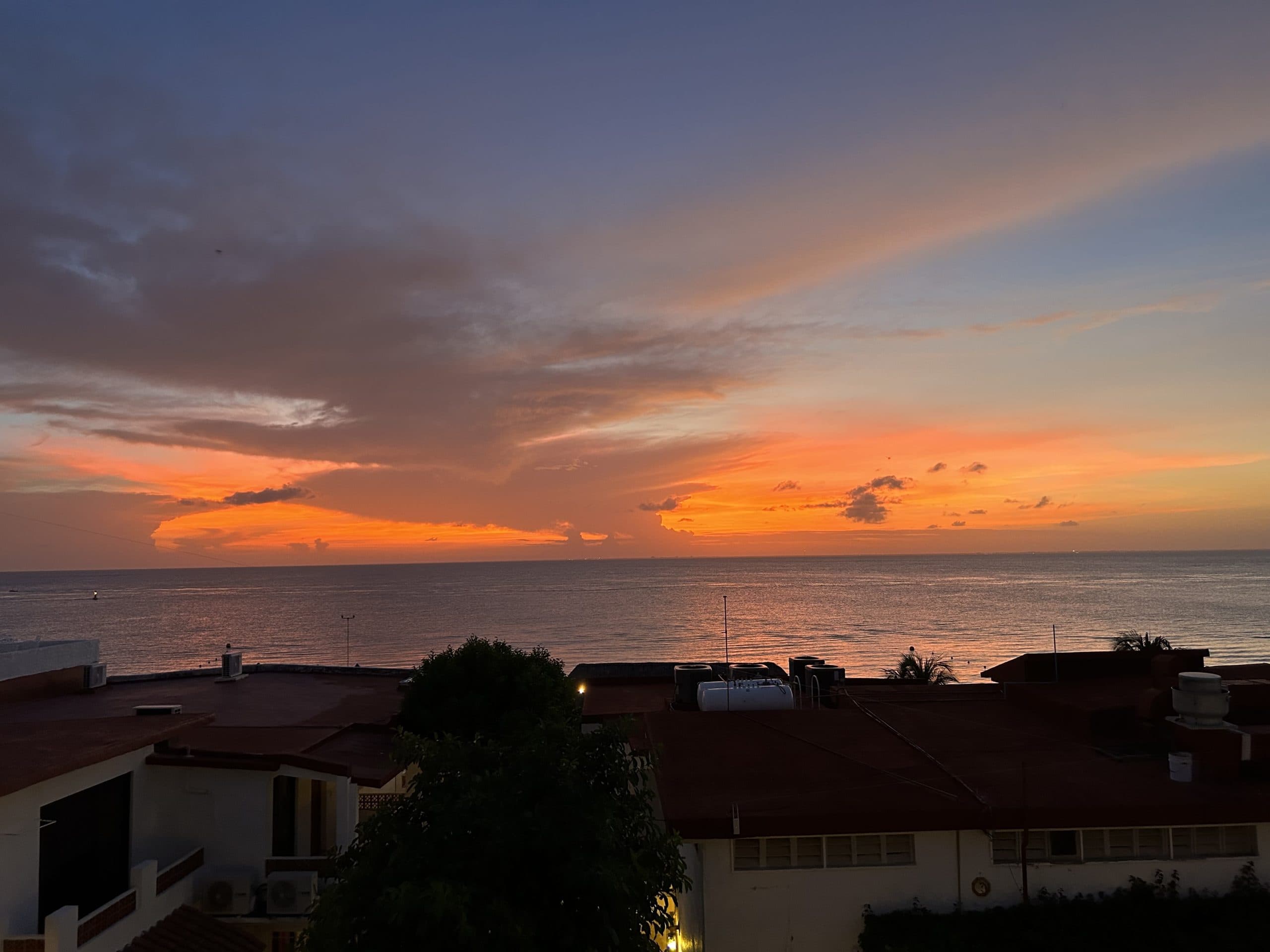 Vibrant sunset over the Caribbean Sea in Cozumel, Mexico, viewed from a rooftop with silhouetted buildings and glowing clouds on the horizon.