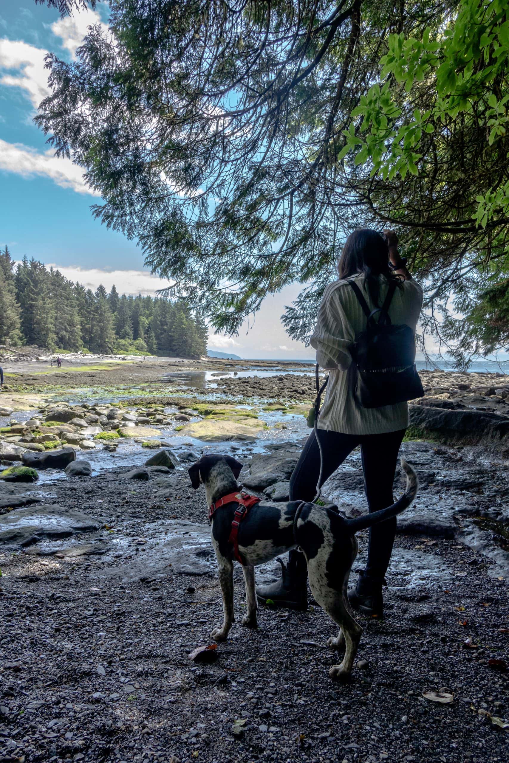Woman with a backpack and her dog enjoying a scenic coastal walk in Port Renfrew, BC, overlooking a rocky shoreline, tidal pools, and forested coastline.