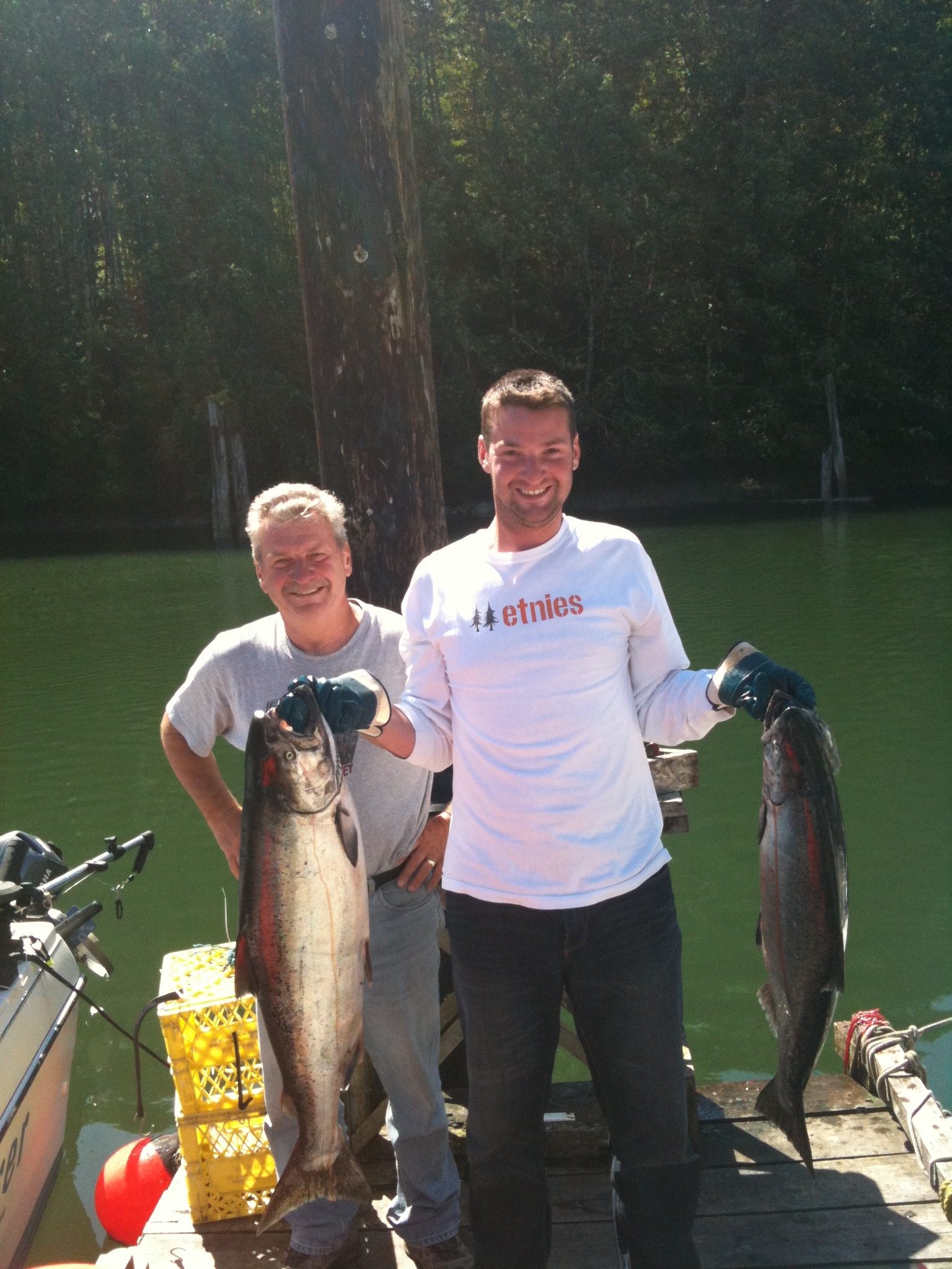 Two smiling anglers standing on a dock in Port Renfrew, BC, proudly holding freshly caught salmon with the marina and forest backdrop.