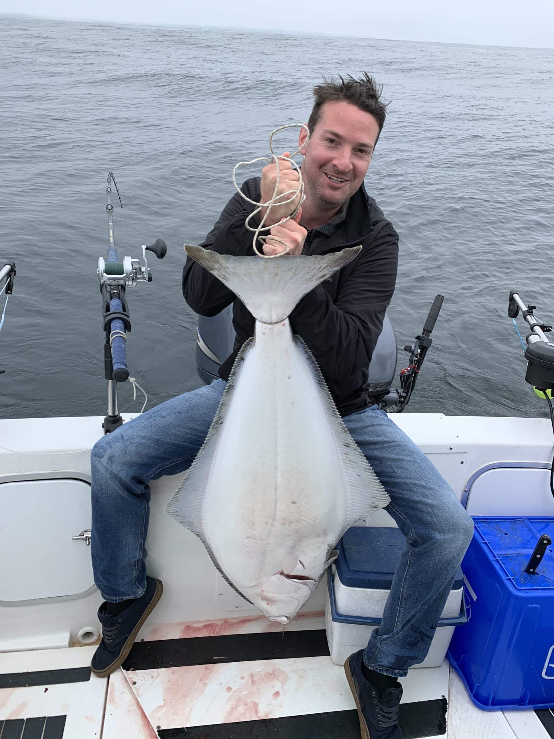 Man on a fishing boat holding a large halibut caught off the coast of Port Renfrew, BC, with the open ocean in the background.
