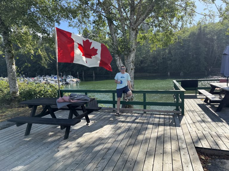 Scenic waterfront view in Port Renfrew, British Columbia with a Canadian flag flying above a wooden dock overlooking a quiet marina surrounded by forested coastline.