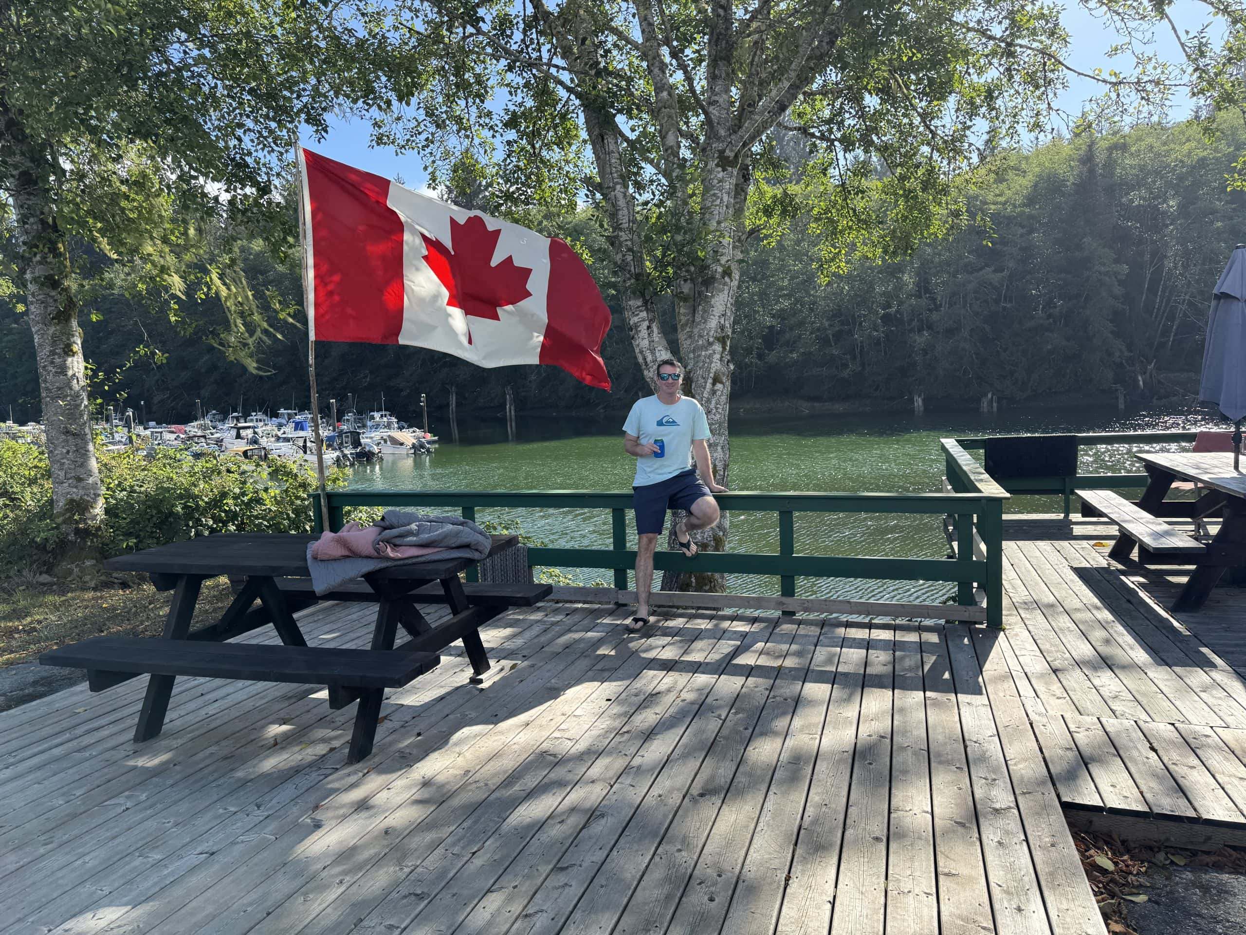 Scenic waterfront view in Port Renfrew, British Columbia with a Canadian flag flying above a wooden dock overlooking a quiet marina surrounded by forested coastline.