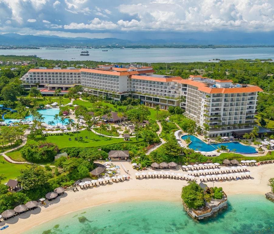 A luxury beachfront resort in Mactan, Cebu, featuring white sand shores, sprawling pools, lush tropical gardens, and stunning views of the sea with the mountains in the distance.