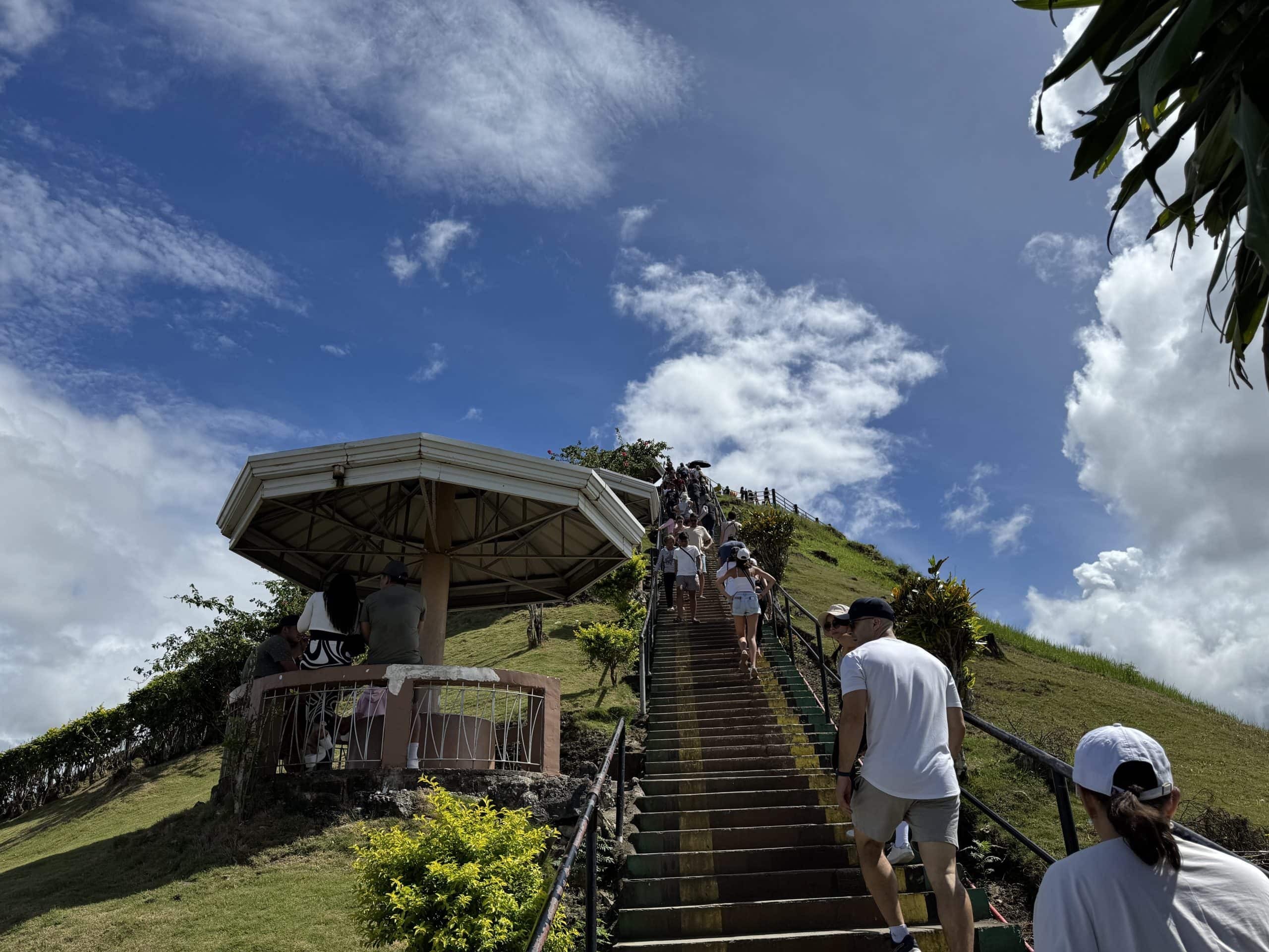 Tourists climbing the steep staircase to the Chocolate Hills viewing deck in Bohol, Philippines under a bright blue sky with scattered clouds