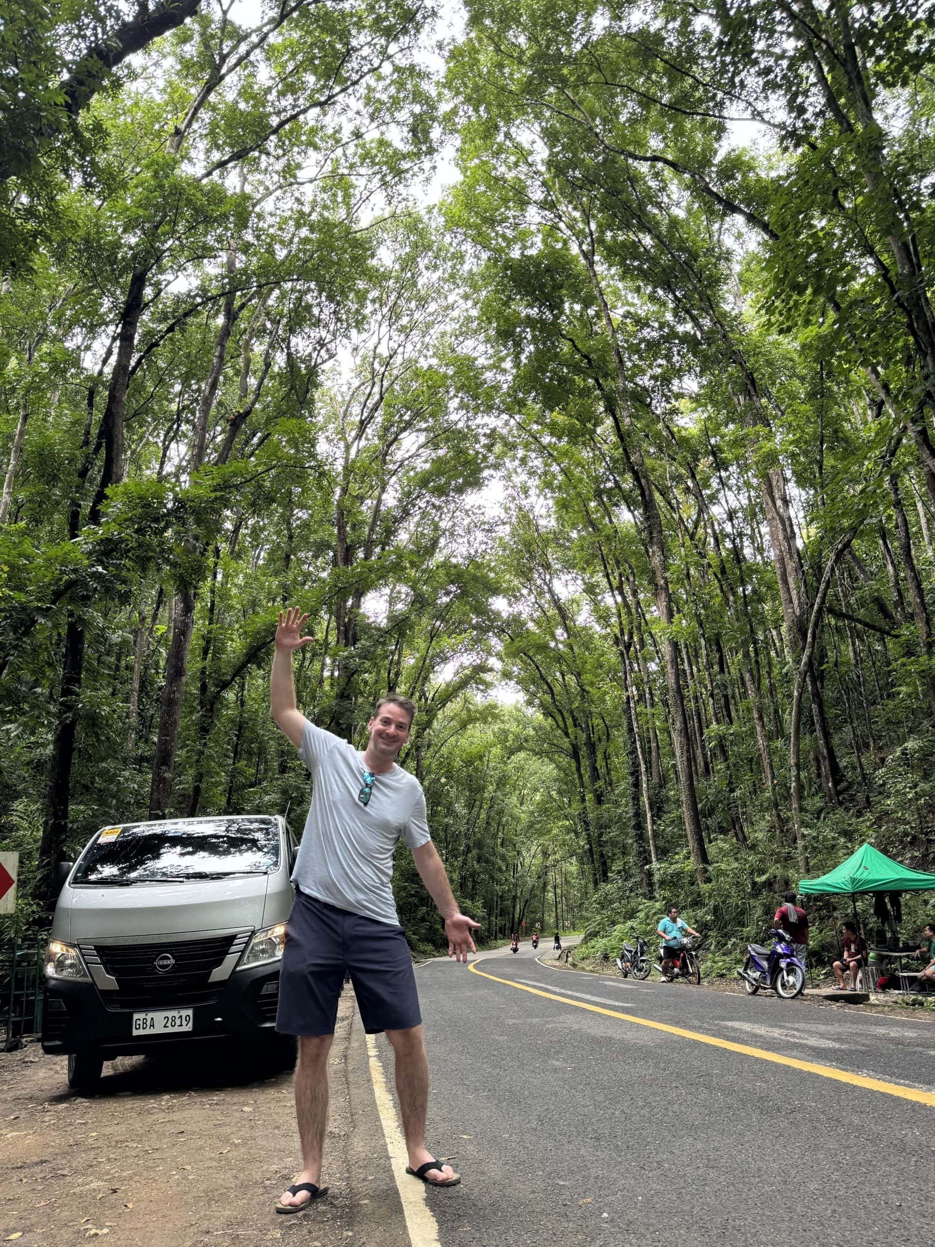 Man posing playfully on the road through the Bilar Man-Made Forest in Bohol, Philippines surrounded by tall mahogany trees forming a scenic green canopy