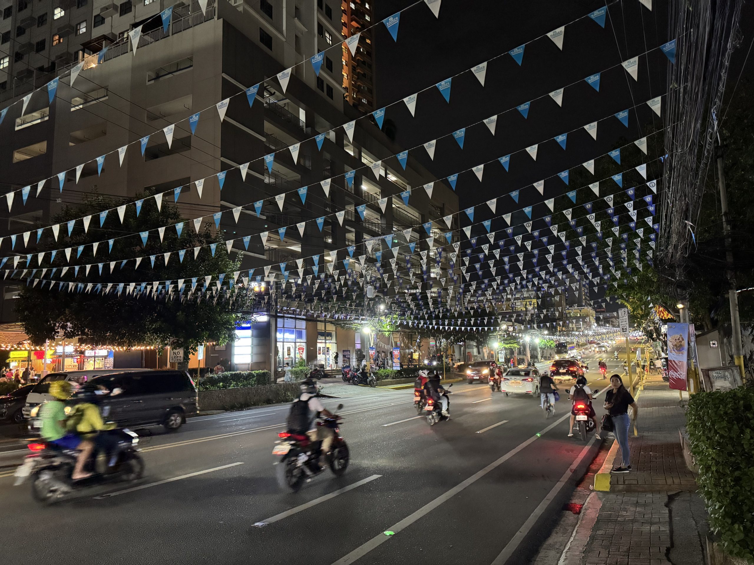 Bustling nightlife along Mango Avenue in Cebu City, with motorcycles, cars, and people under festive blue and white flags that highlight the energy of this famous entertainment street.