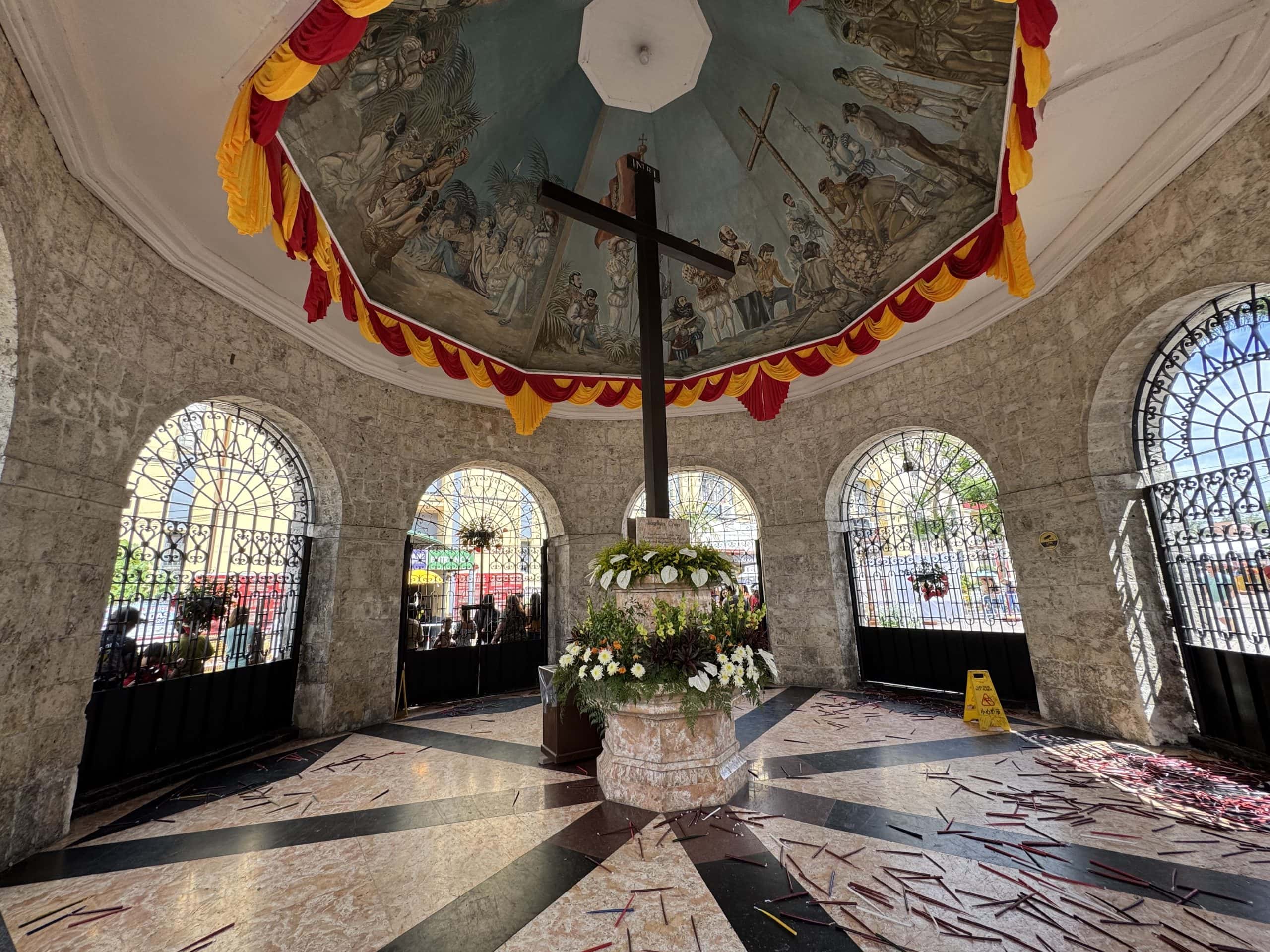 The iconic Magellan’s Cross in Cebu City, housed in a stone chapel with painted ceiling murals and decorated with flowers, a historic landmark central to the city’s culture and faith.