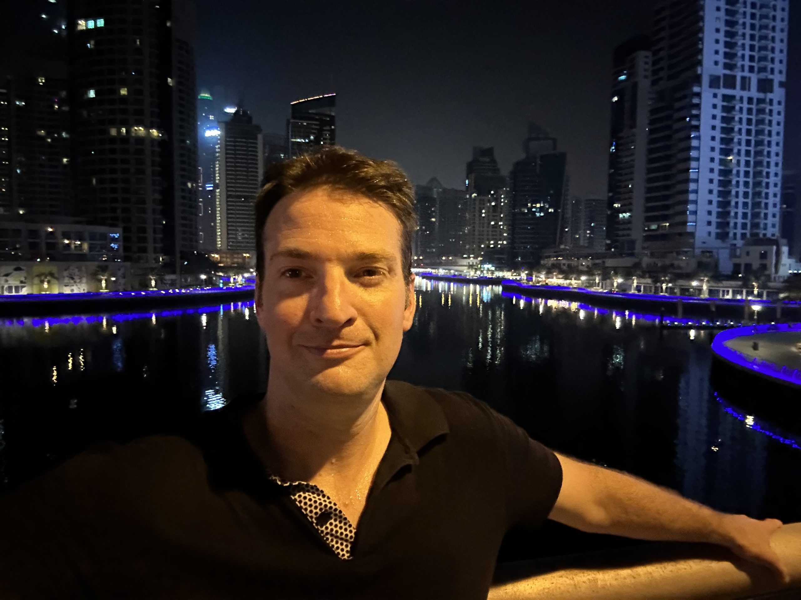 Man enjoying a night in Dubai Marina with the city’s glittering skyline reflected on the calm water and vibrant blue lights along the promenade.