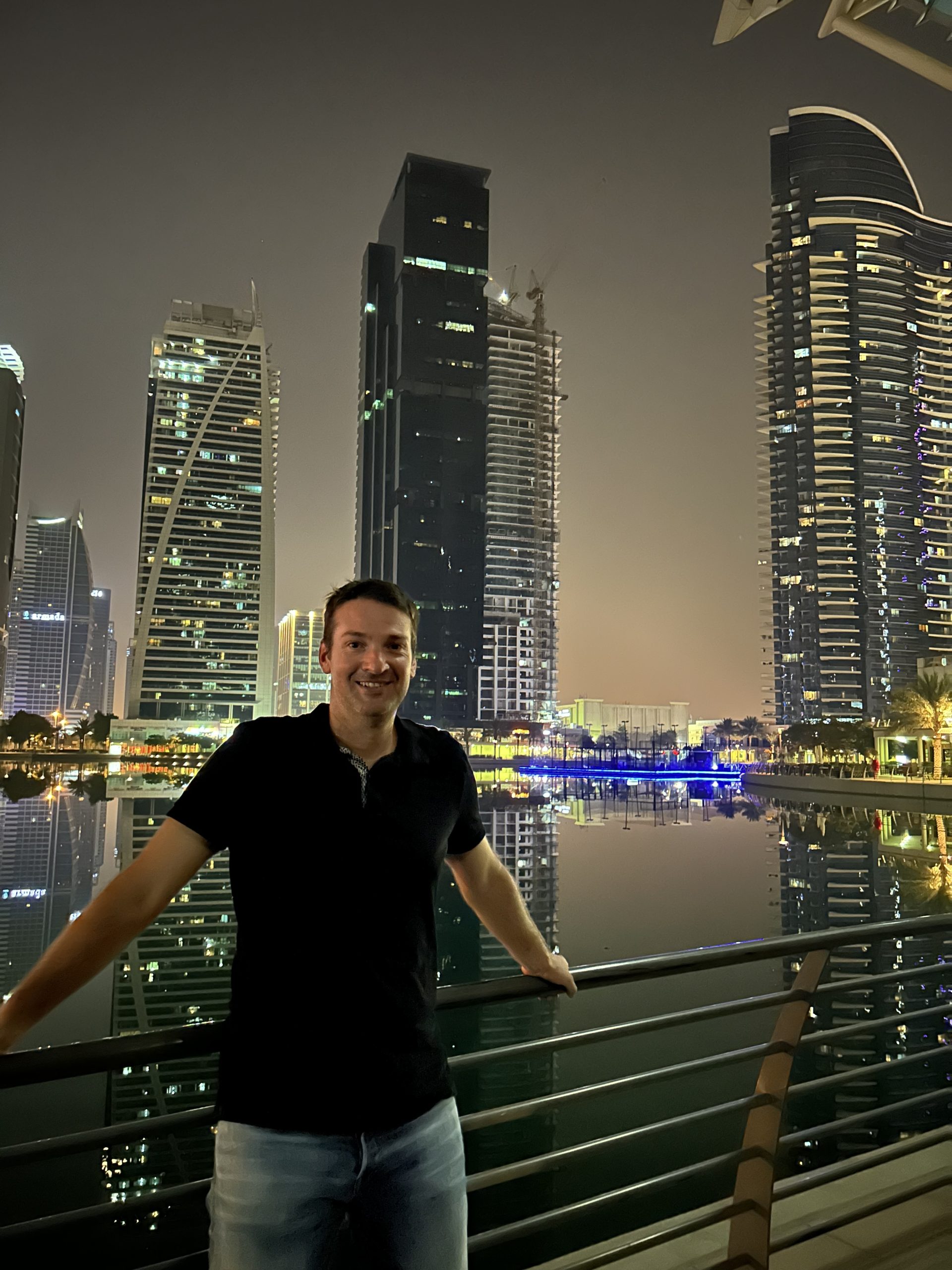 Man standing by the waterfront in Dubai at night with modern skyscrapers reflecting on the calm water, showcasing the city’s vibrant skyline and urban atmosphere.