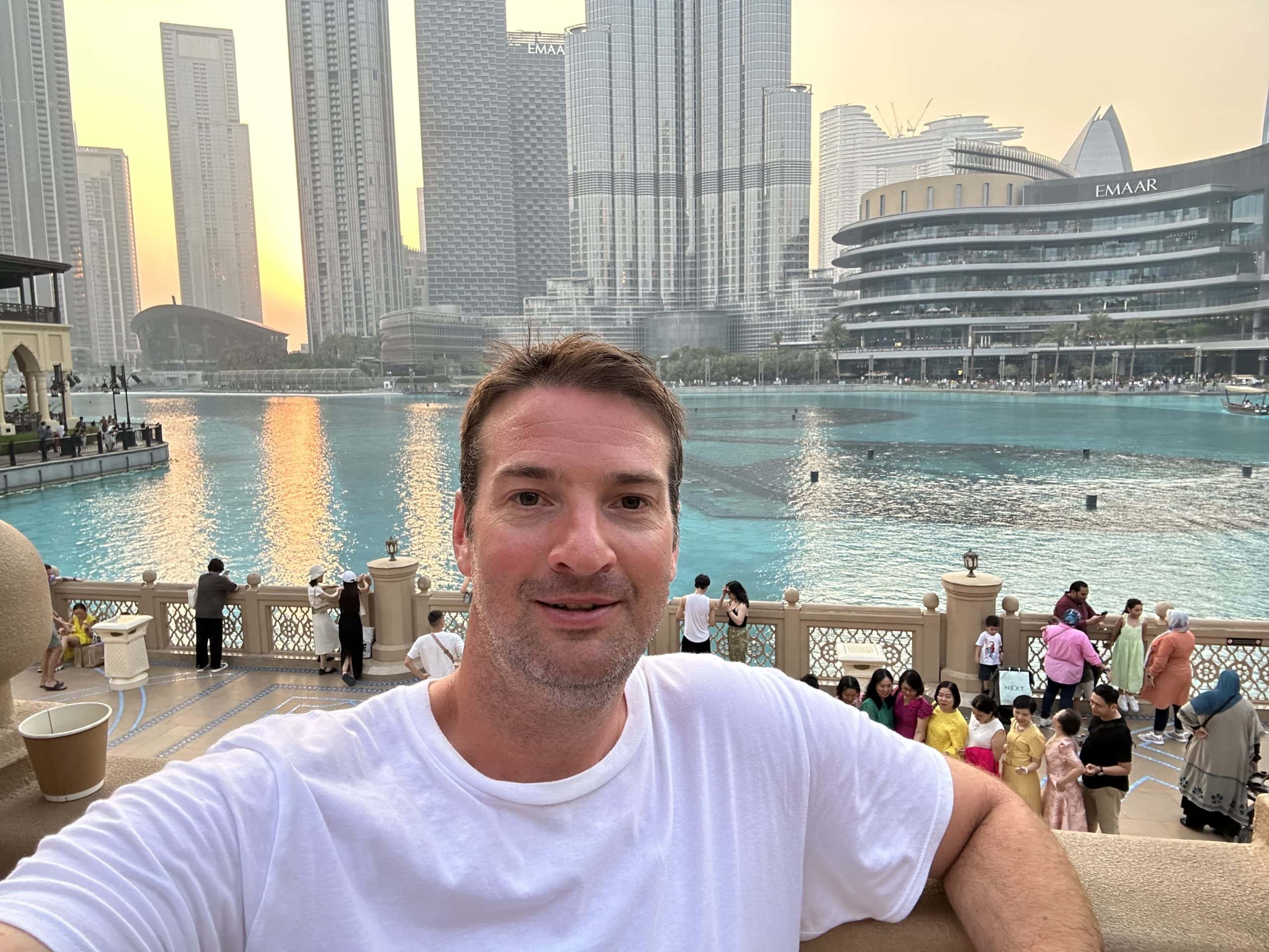Traveler taking a selfie at the Dubai Fountain during sunset, with turquoise water, modern skyscrapers, and the iconic Dubai Mall skyline in the background.