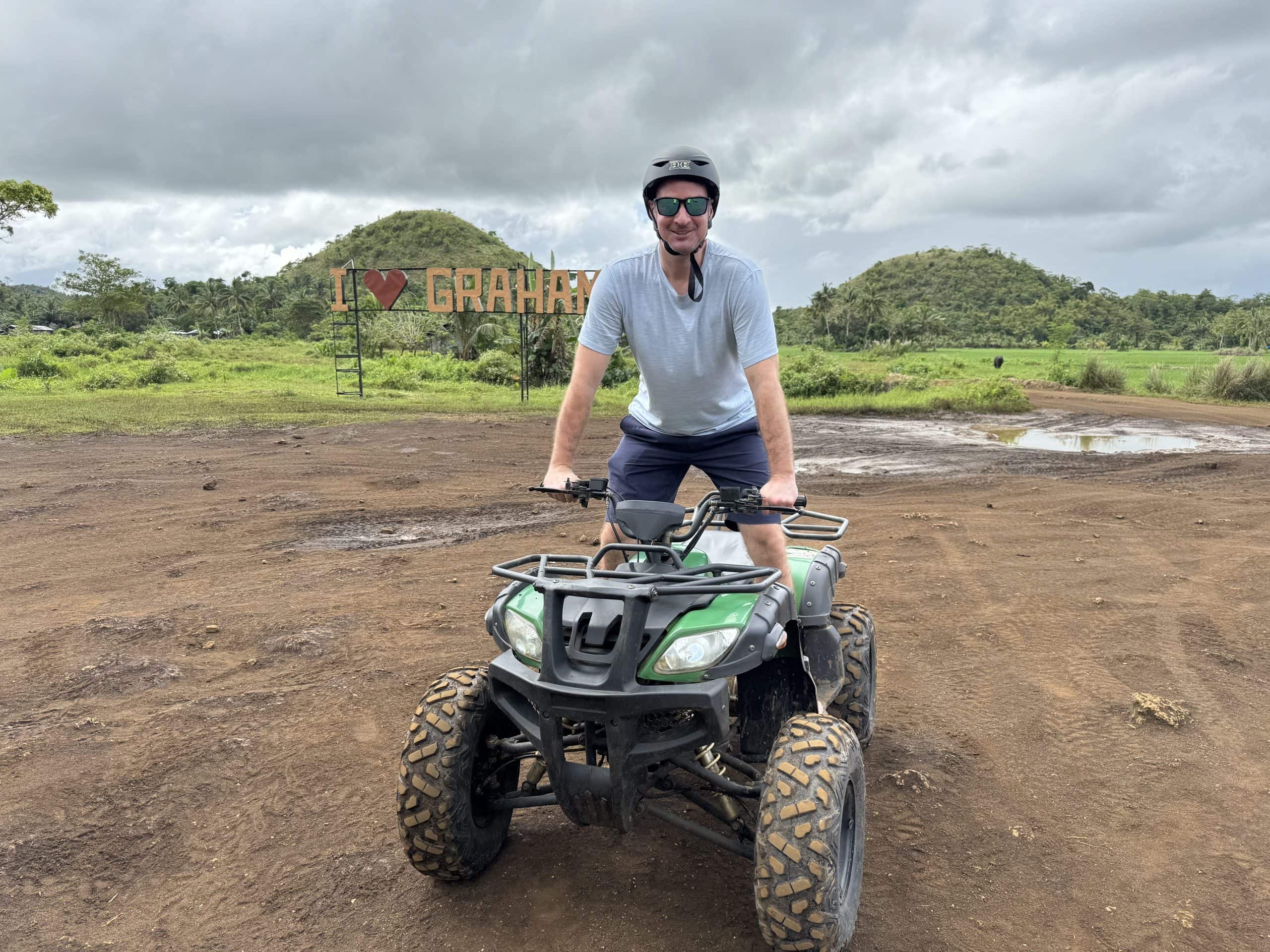 Man riding an ATV on a dirt trail near the Chocolate Hills in Bohol, Philippines with green hills and cloudy skies in the background