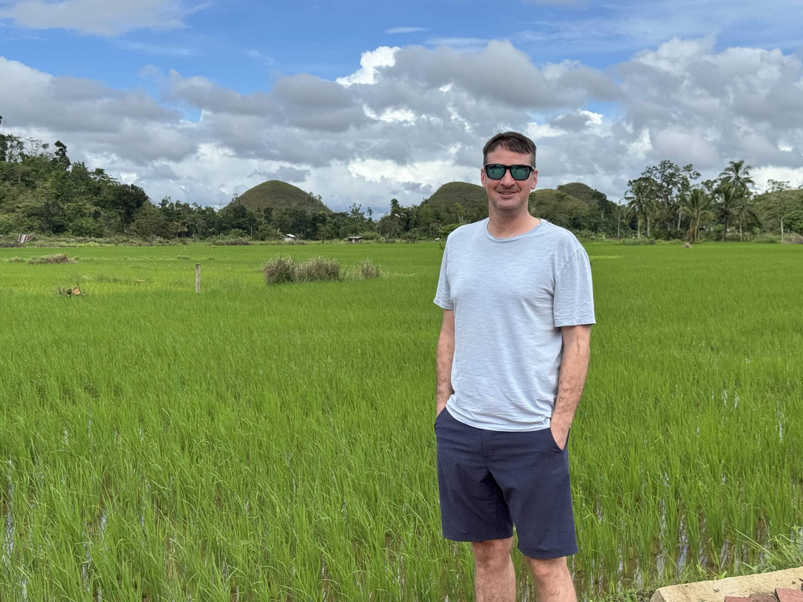 Man standing in front of lush green rice fields with the Chocolate Hills in the background in Bohol, Philippines on a partly cloudy day