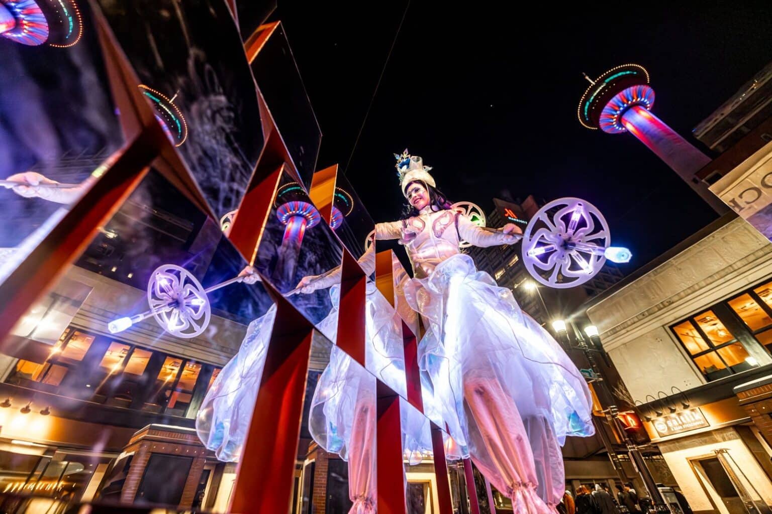 A vibrant performer on stilts dazzles the crowd with glowing props during Chinook Blast in Calgary, as the Calgary Tower lights up the night sky in the background.