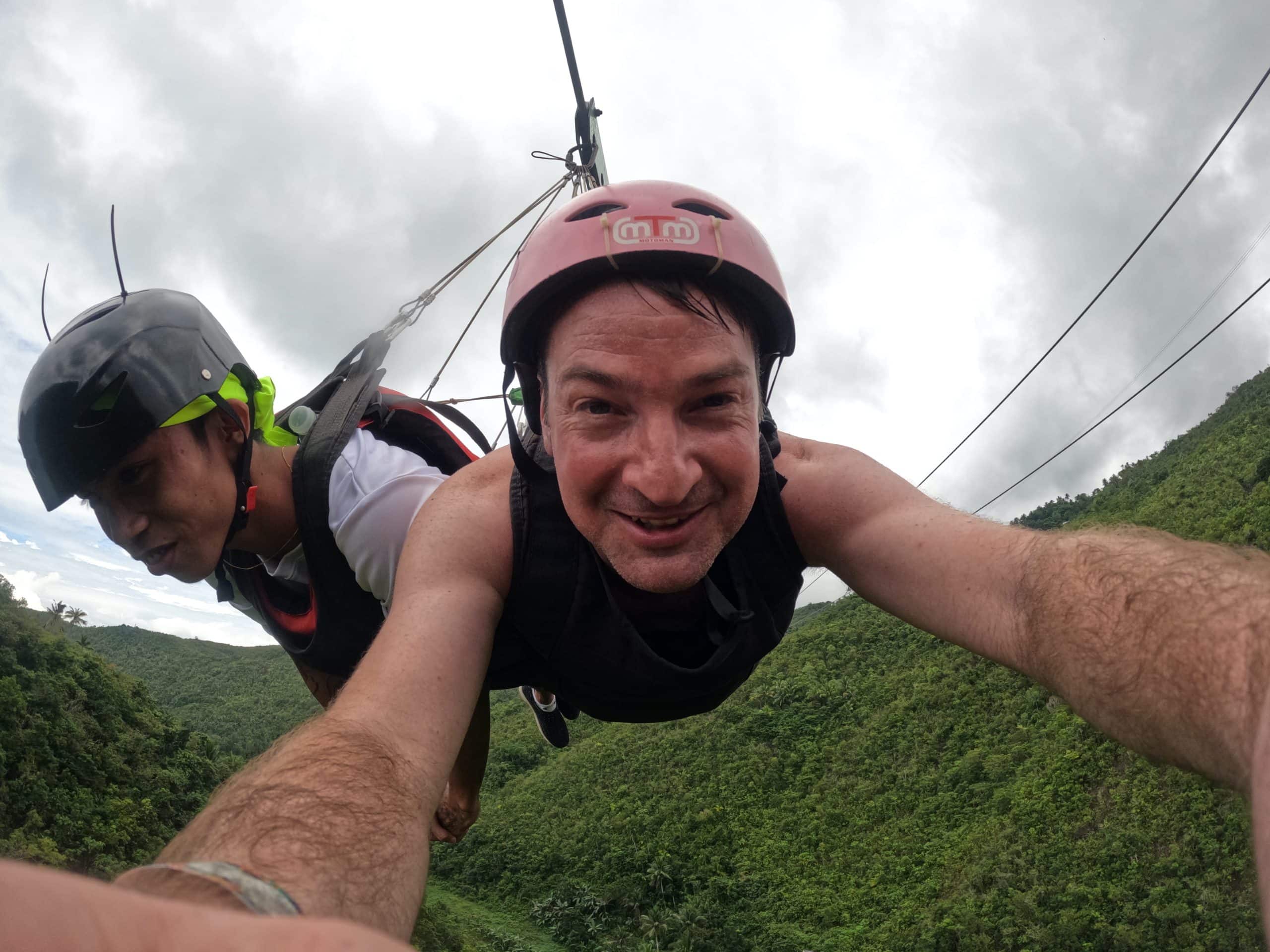 A traveller soars over lush green mountains on a zipline adventure in Kawasan Falls Cebu, capturing the thrill of the ride high above the forest canopy.