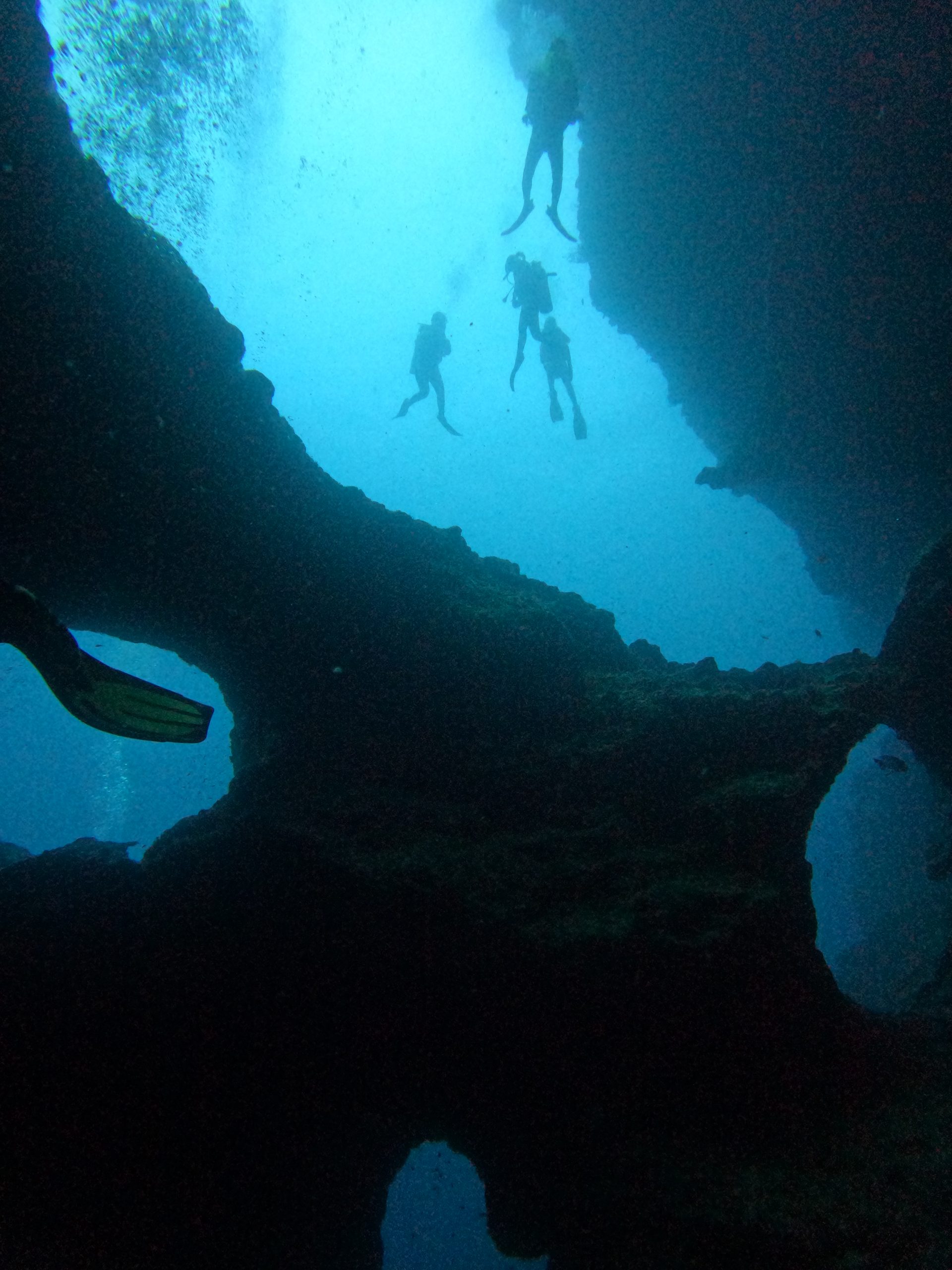 Scuba divers explore the dramatic underwater rock formations and archways of Pescador Island in Moalboal, surrounded by deep blue water and schools of fish.