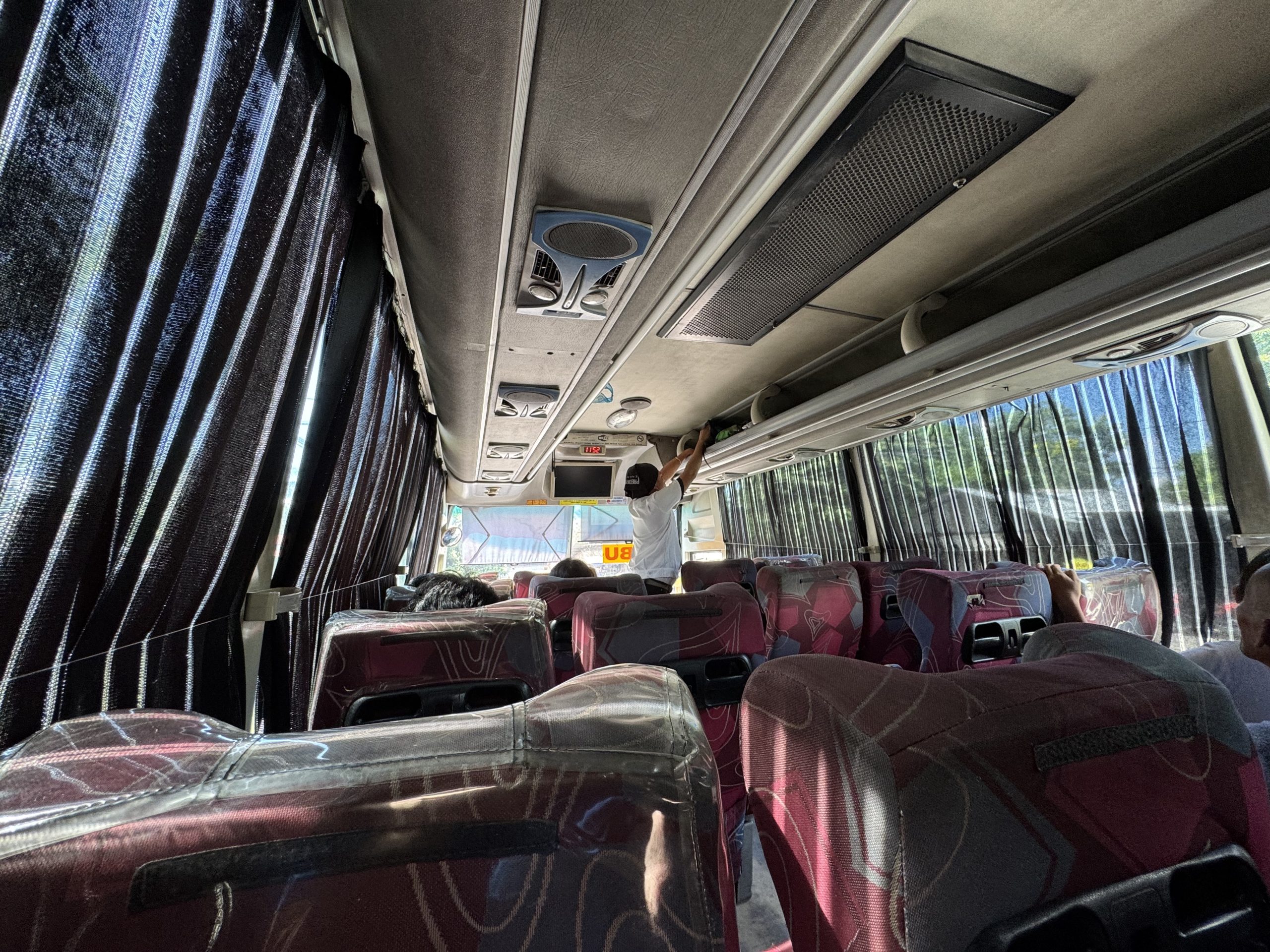 Inside a Ceres bus traveling through Cebu, sunlight filters through dark curtains as passengers settle into red patterned seats on the journey toward Moalboal.