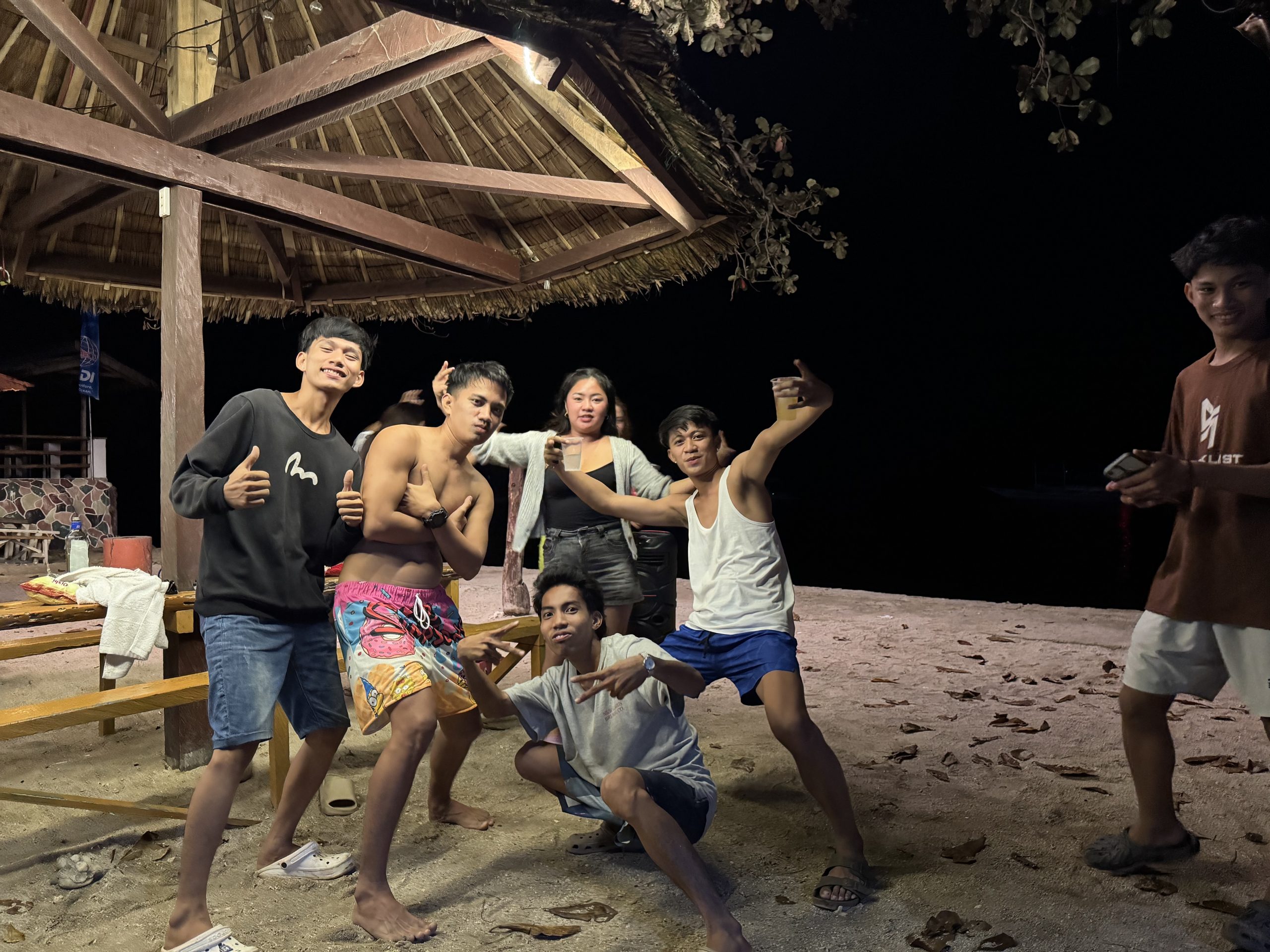 A lively group of friends hangs out under a beach hut in Moalboal at night, enjoying music and drinks by the shore with a relaxed island vibe.