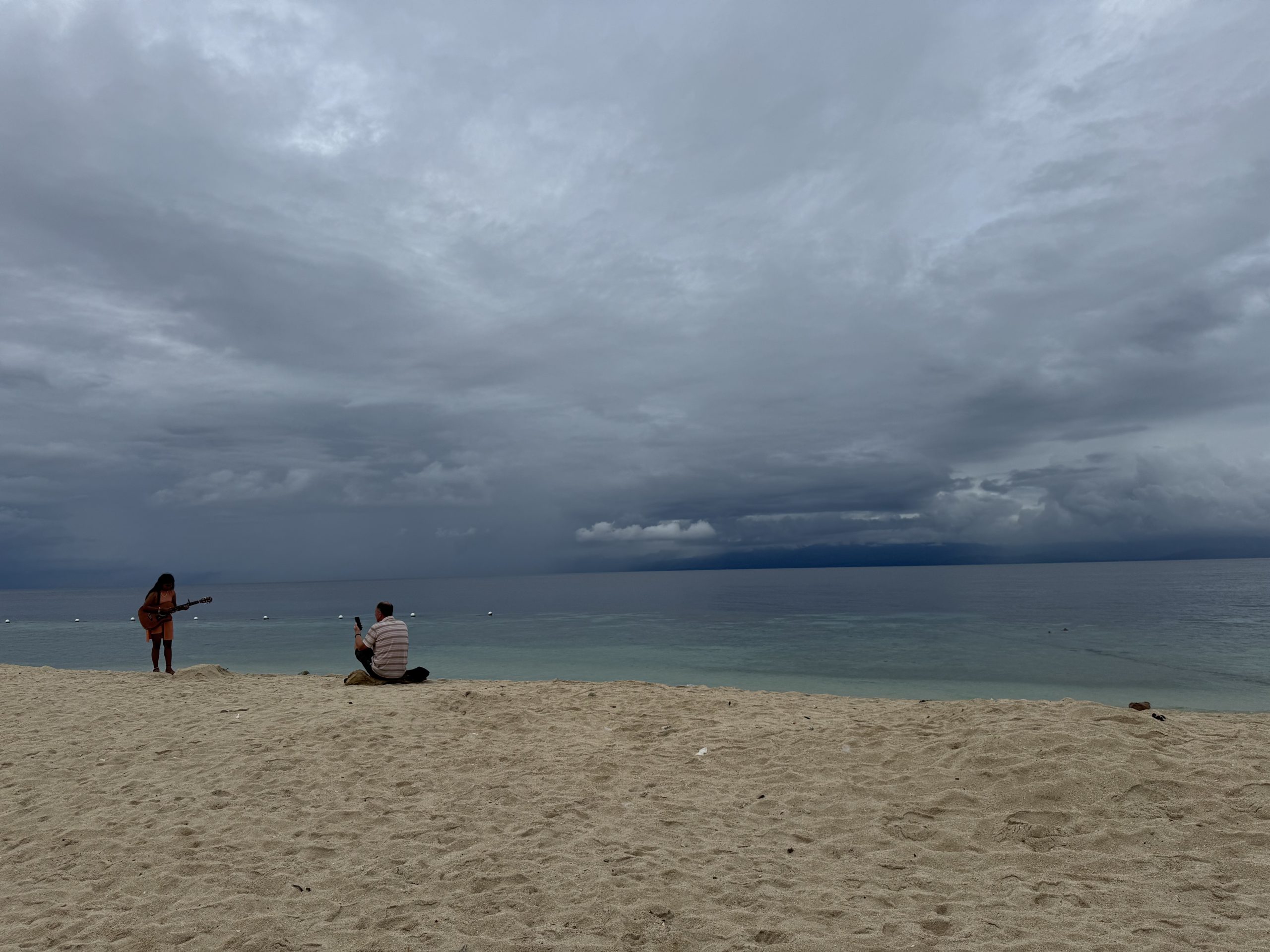 A quiet moment on White Beach in Moalboal, Cebu, as a musician plays guitar by the shore under an overcast sky with calm turquoise waters in the background.