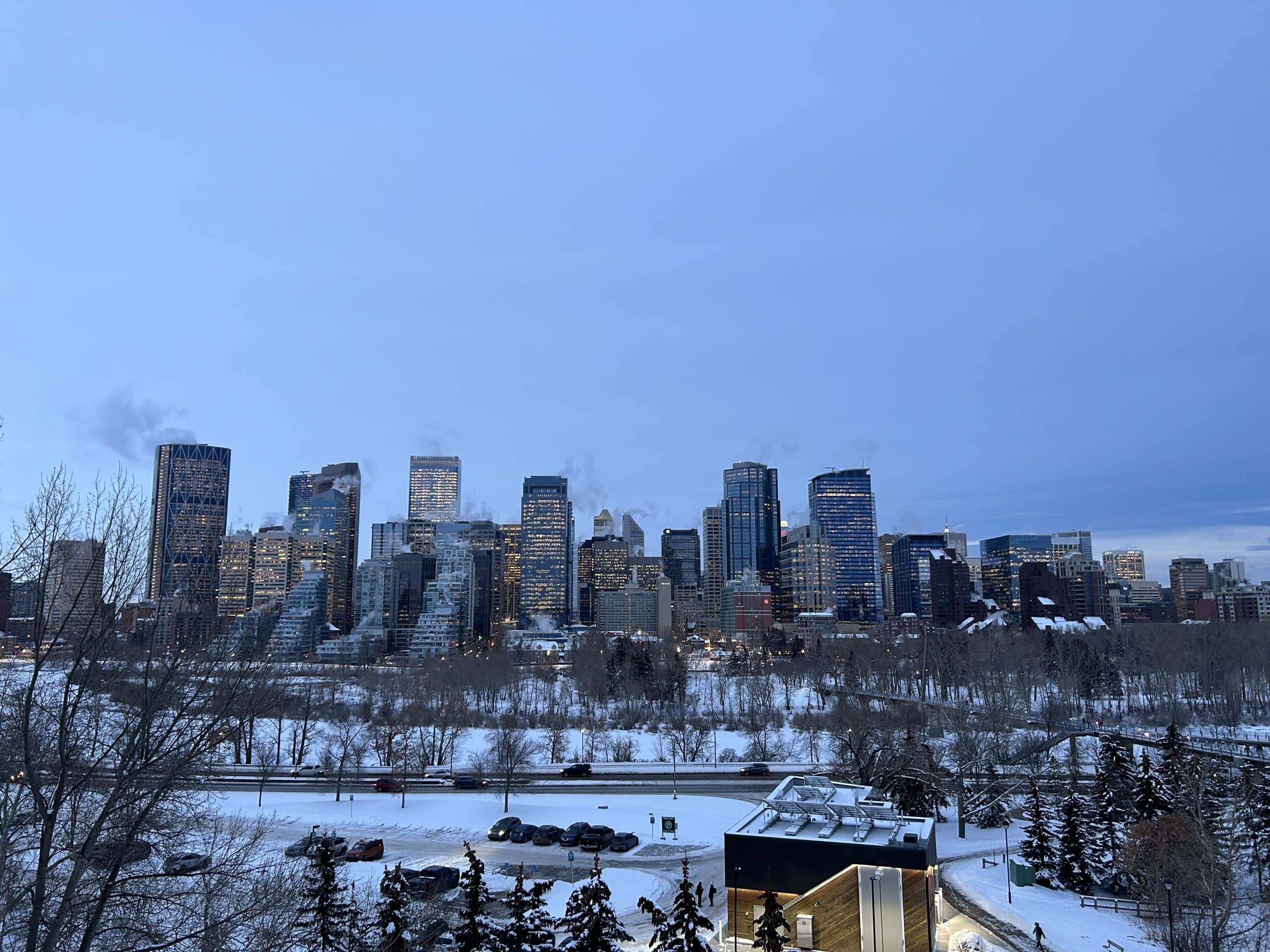 Downtown Calgary glows at dusk with snow-covered parks and trees in the foreground, showcasing the city’s winter skyline under a peaceful blue evening sky.