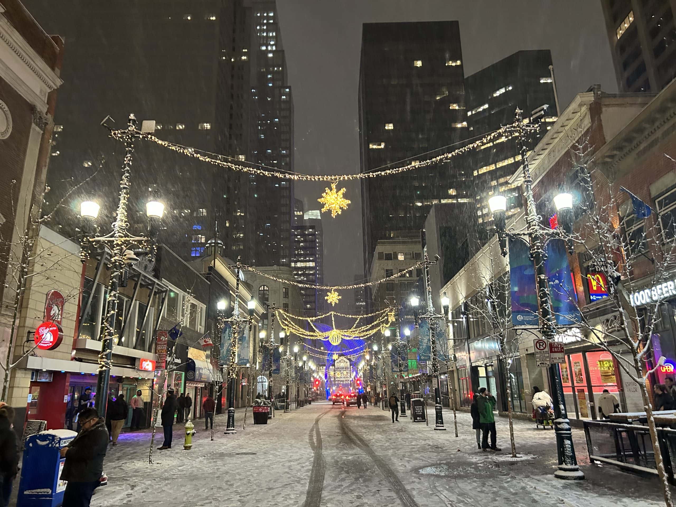 Snow falls over a brightly lit Stephen Ave in Calgary decorated with golden holiday lights and festive displays, as people stroll past shops and restaurants in the evening.