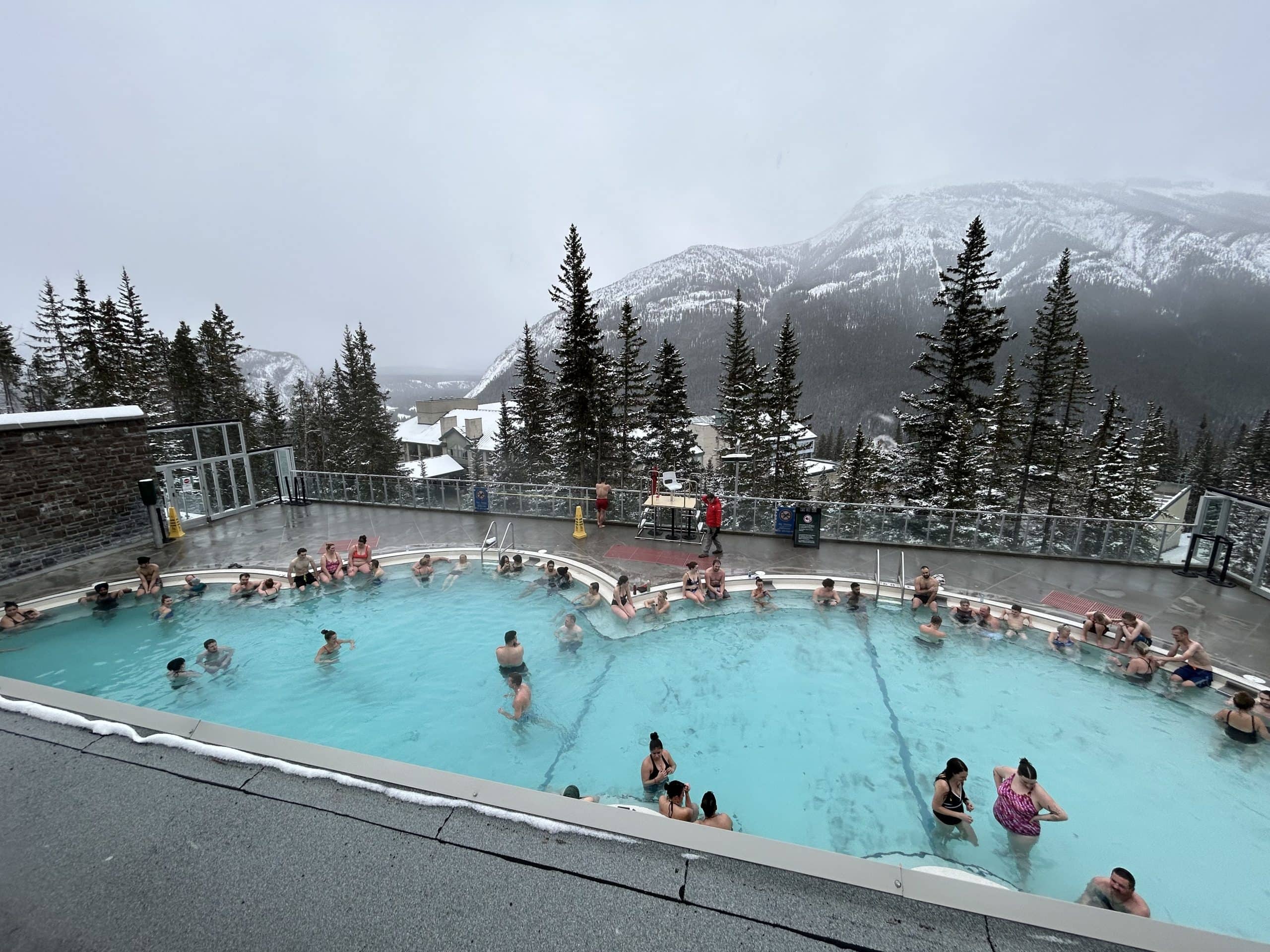 Visitors relax in the steaming Banff Upper Hot Springs, surrounded by snowy pine trees and breathtaking mountain views on a chilly winter day in the Canadian Rockies.