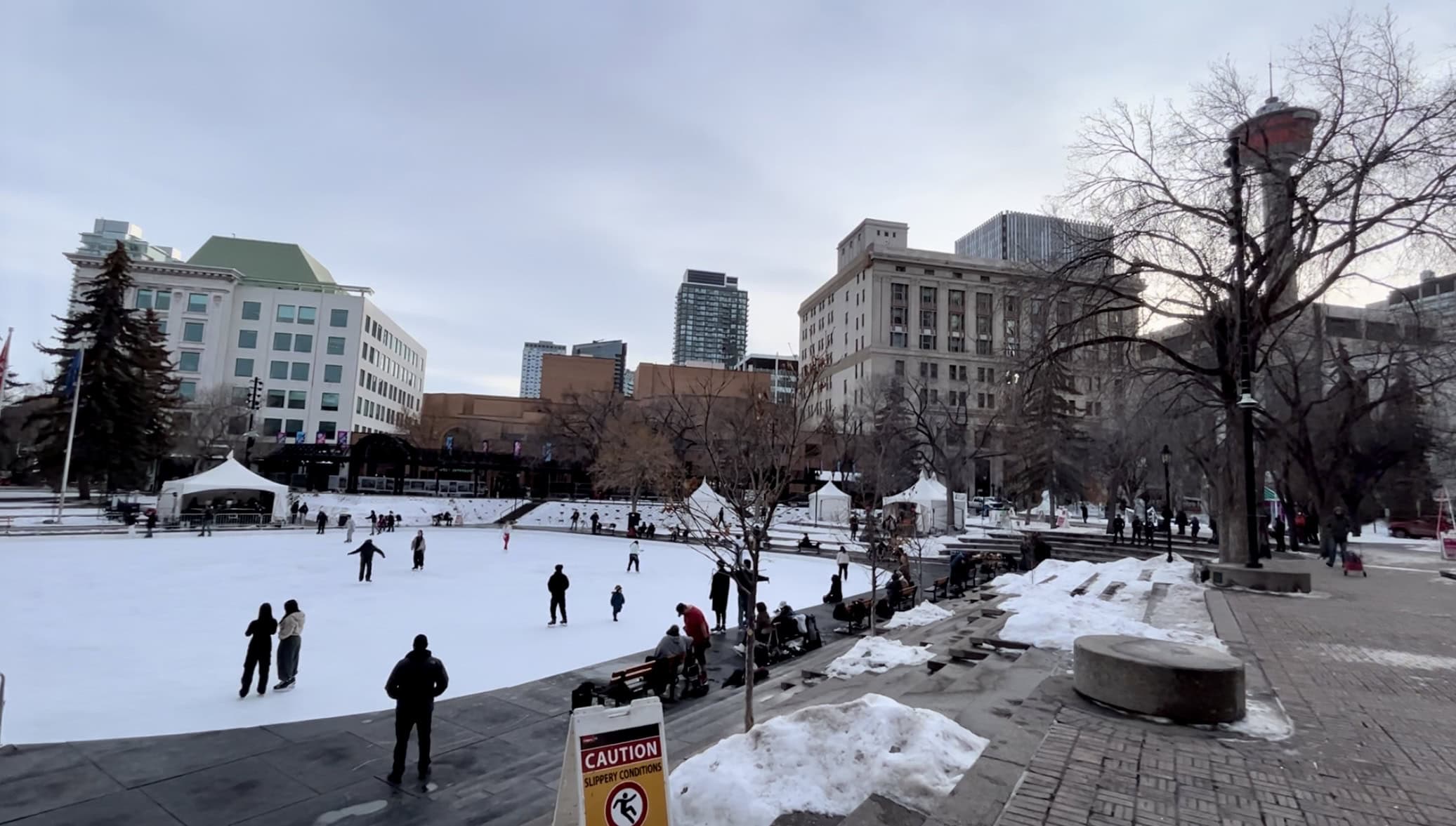 Skaters enjoy the outdoor ice rink at Olympic Plaza in downtown Calgary, with historic buildings, modern towers, and the Calgary Tower creating a scenic winter backdrop.
