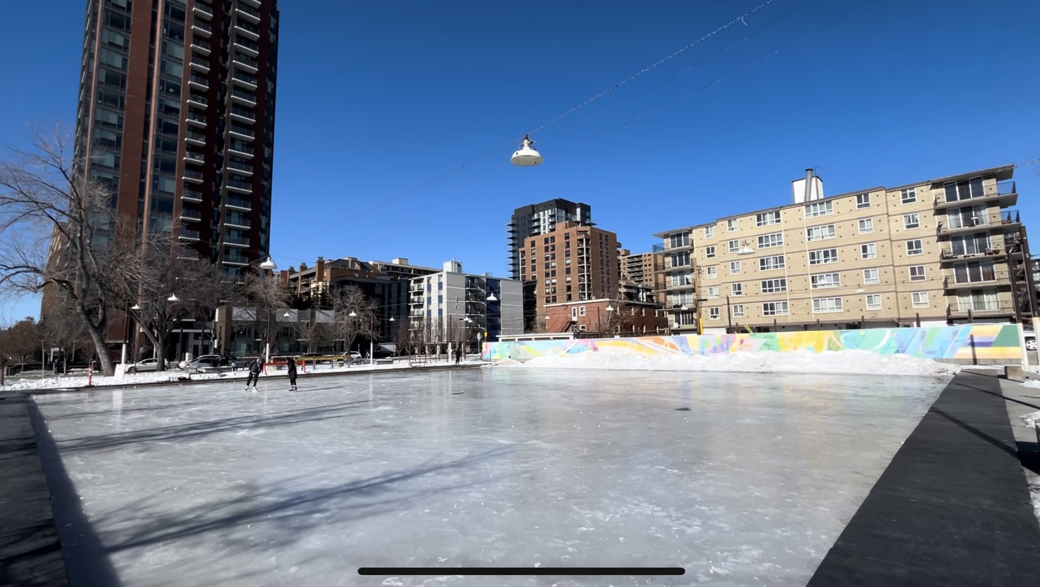 Skaters glide across the outdoor ice rink at Thomson Family Park in Calgary, surrounded by colourful murals and city buildings under a clear blue winter sky.