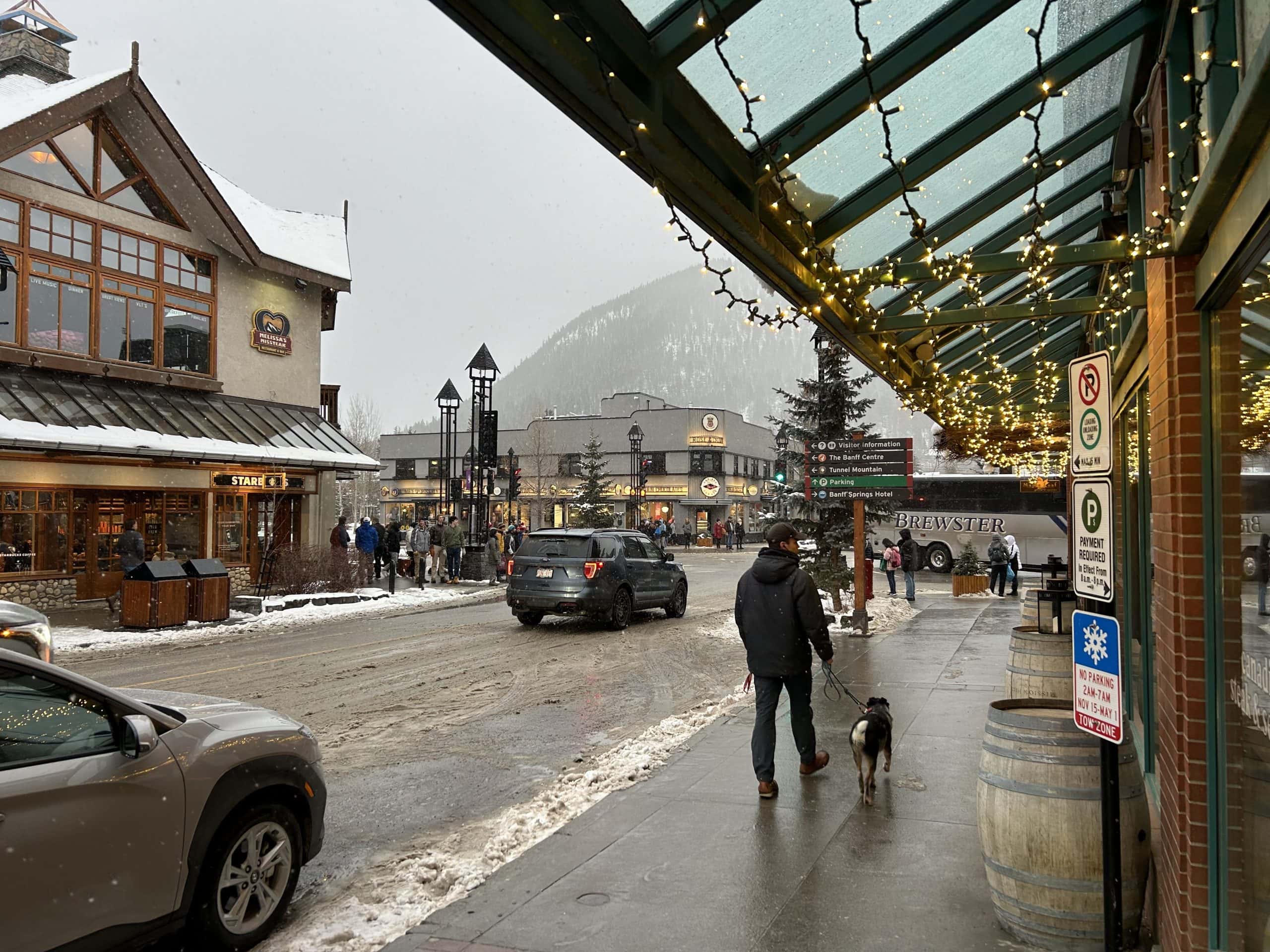 Snow falls over Banff Avenue as people stroll past cozy shops and restaurants, with festive lights, mountain views, and a lively winter atmosphere in the heart of Banff.