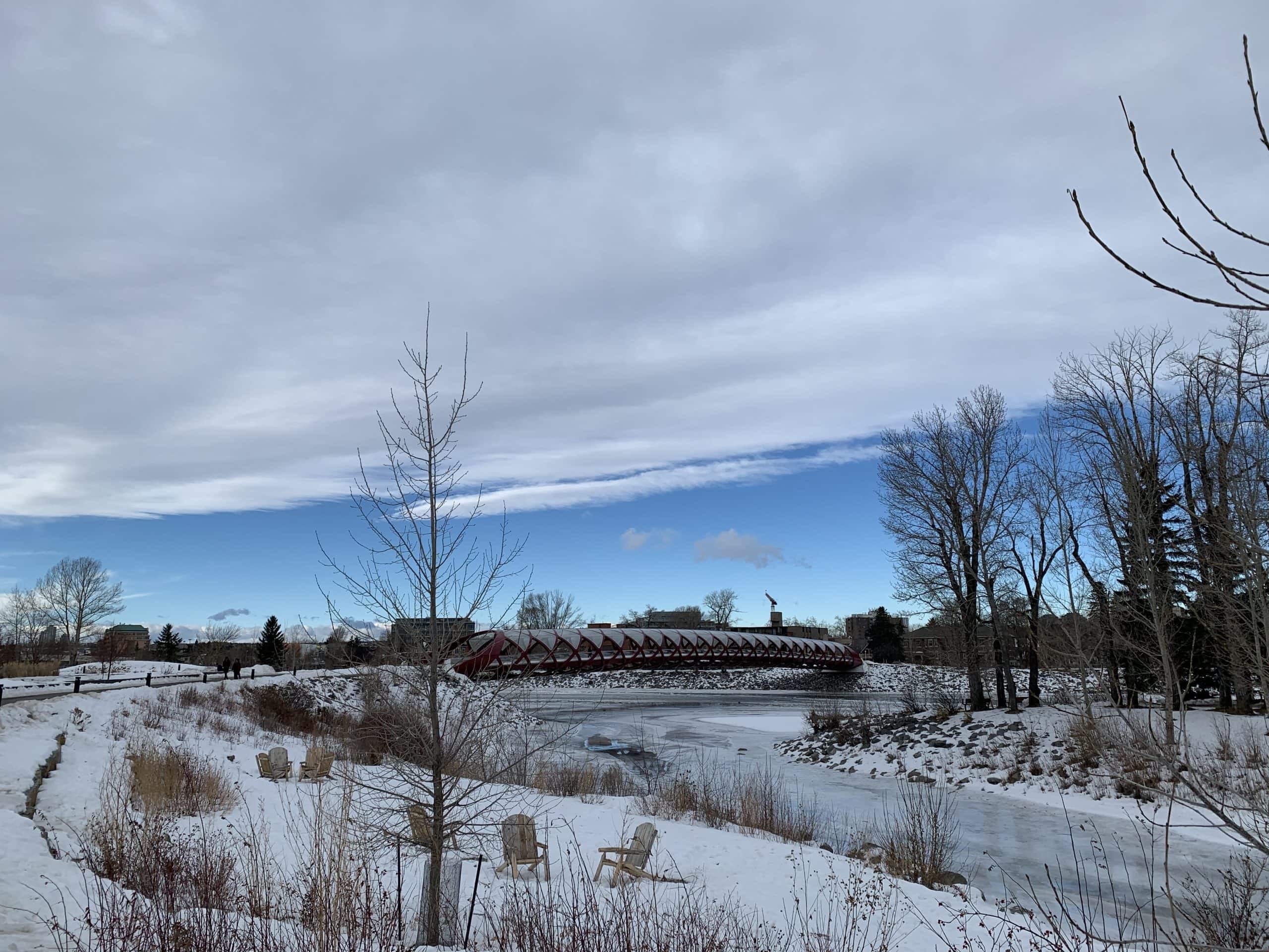 The Peace Bridge spans the partially frozen Bow River in Calgary, surrounded by snowy riverbanks, bare winter trees, and a crisp blue sky breaking through the clouds.