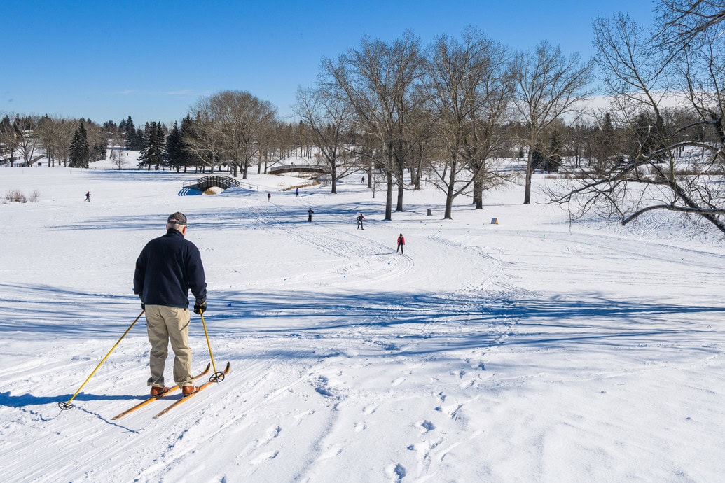 Cross-country skiers enjoy the groomed snowy trails at Confederation Park in Calgary, surrounded by open fields, leafless trees, and bright blue winter skies.