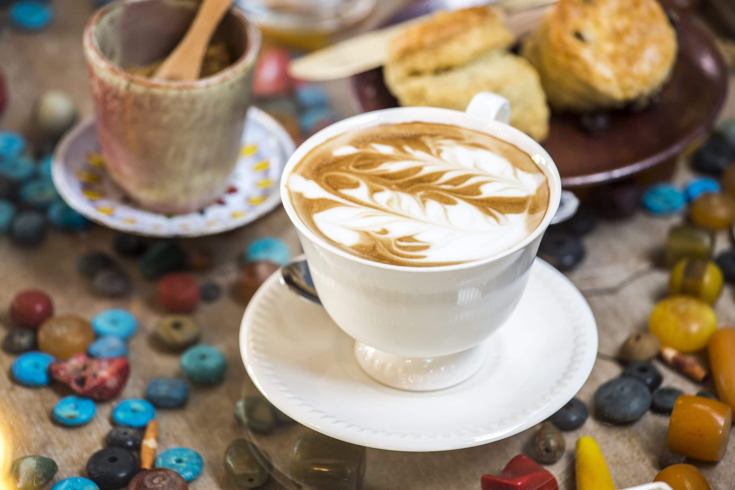 A beautifully crafted latte with intricate foam art sits in a white cup and saucer, surrounded by colorful decor and paired with fresh pastries for a cozy café experience.