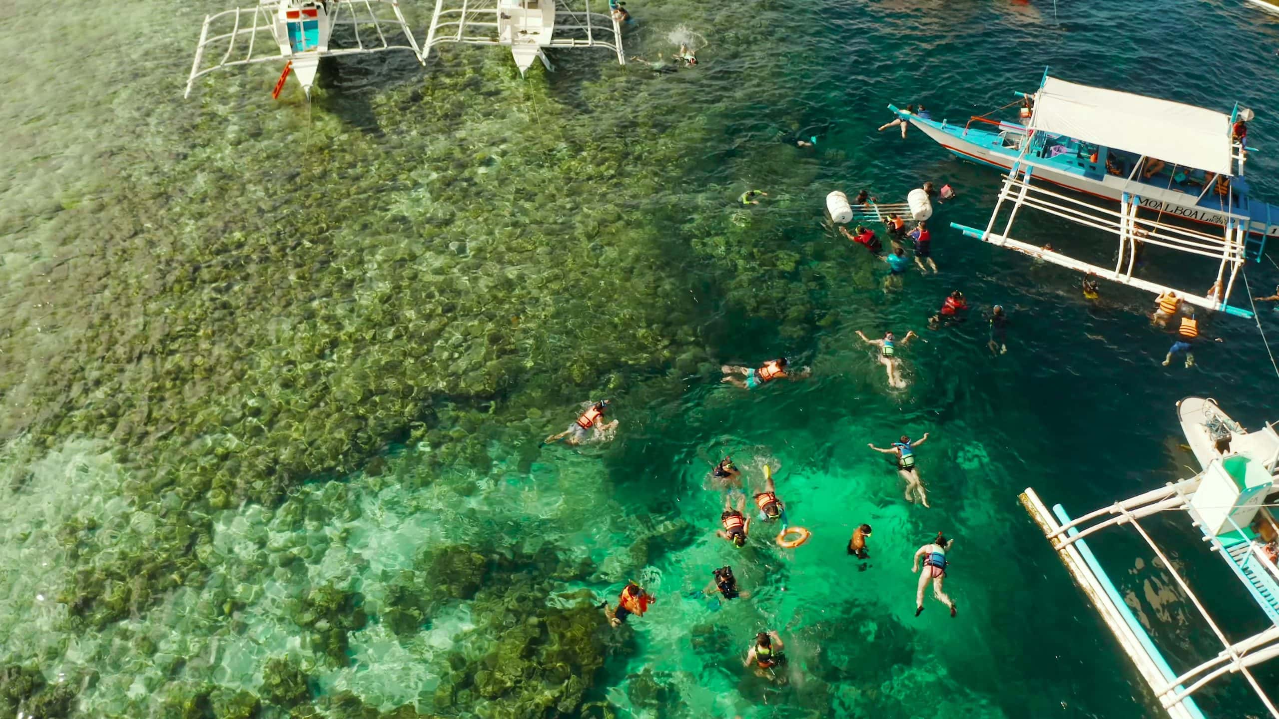Tourists snorkel in the crystal-clear turquoise waters of Moalboal, Cebu, surrounded by coral reefs and traditional outrigger boats near Panagsama Beach.
