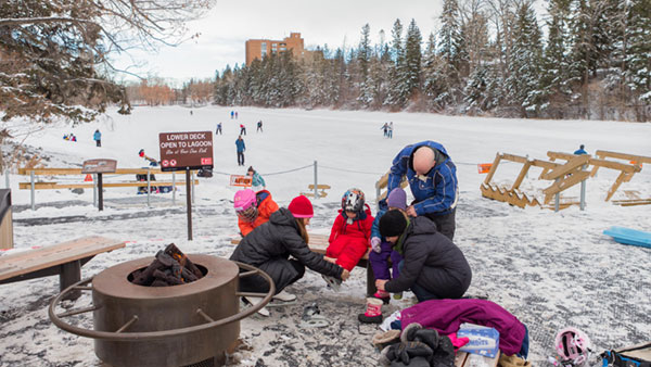 Families lace up their skates beside a fire pit at Bowness Park in Calgary, while skaters enjoy the frozen lagoon surrounded by snowy trees and winter scenery.