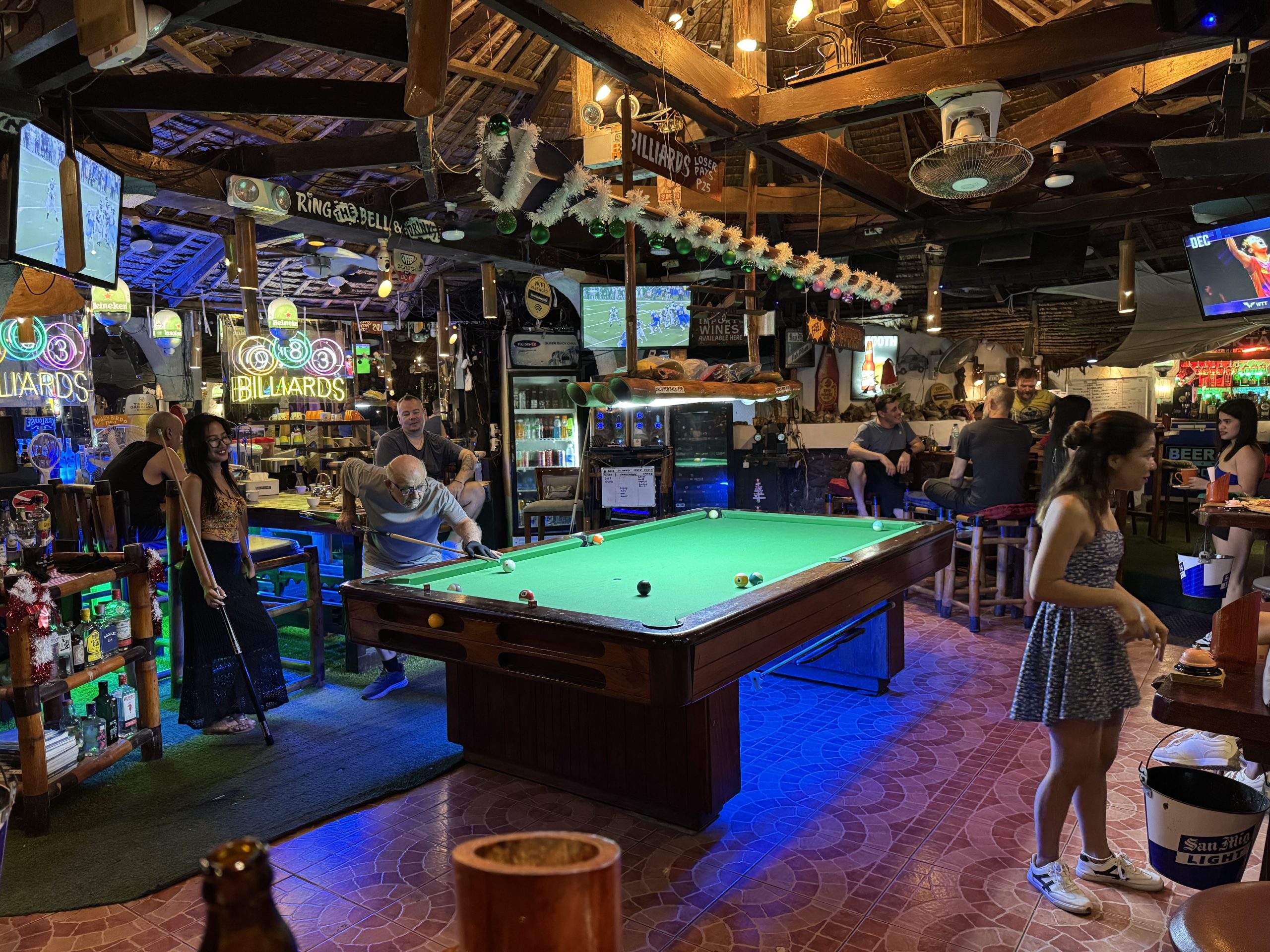 Inside Big Apple Dive Resort in Sabang, Puerto Galera, showing divers and travellers playing pool, watching sports, and relaxing in the lively bar area after a day of scuba diving.