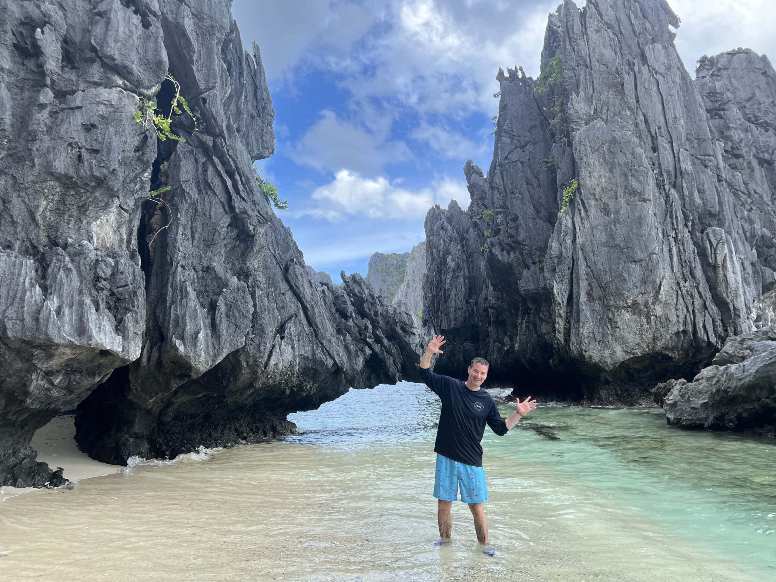 A traveler standing in shallow turquoise water between dramatic limestone cliffs on a hidden beach in El Nido, Palawan, Philippines.