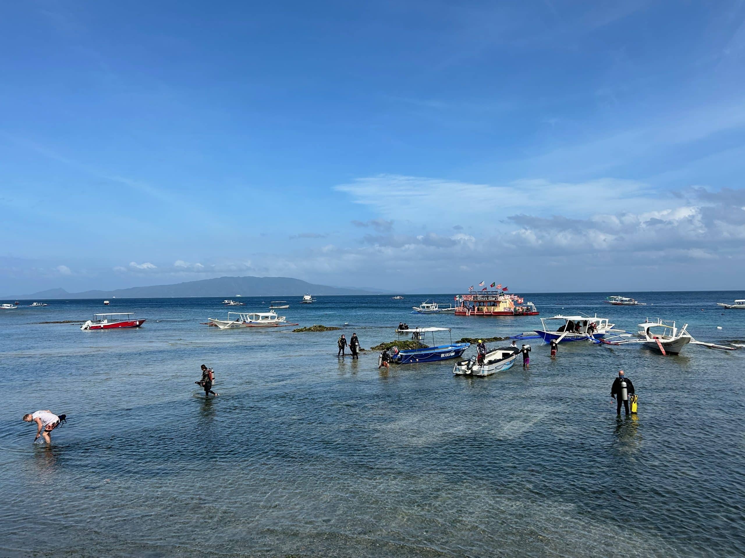 Scuba divers preparing to enter the water in Sabang, Puerto Galera, Philippines, with dive boats anchored offshore.