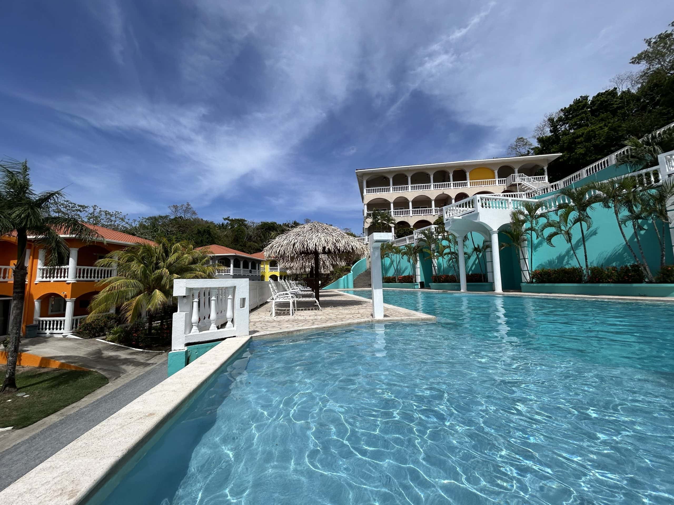 Resort pool in Roatán, Honduras with turquoise water, palm trees, and colourful Caribbean style buildings on a sunny day.
